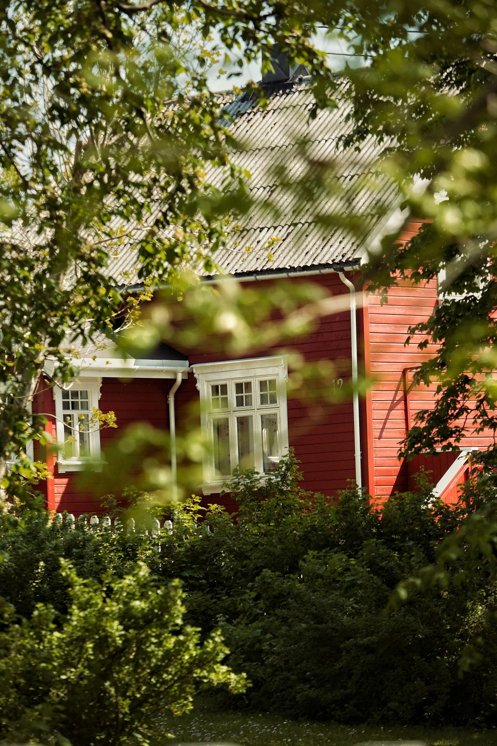 A red wooden house with white-framed windows, surrounded by green trees and bushes.