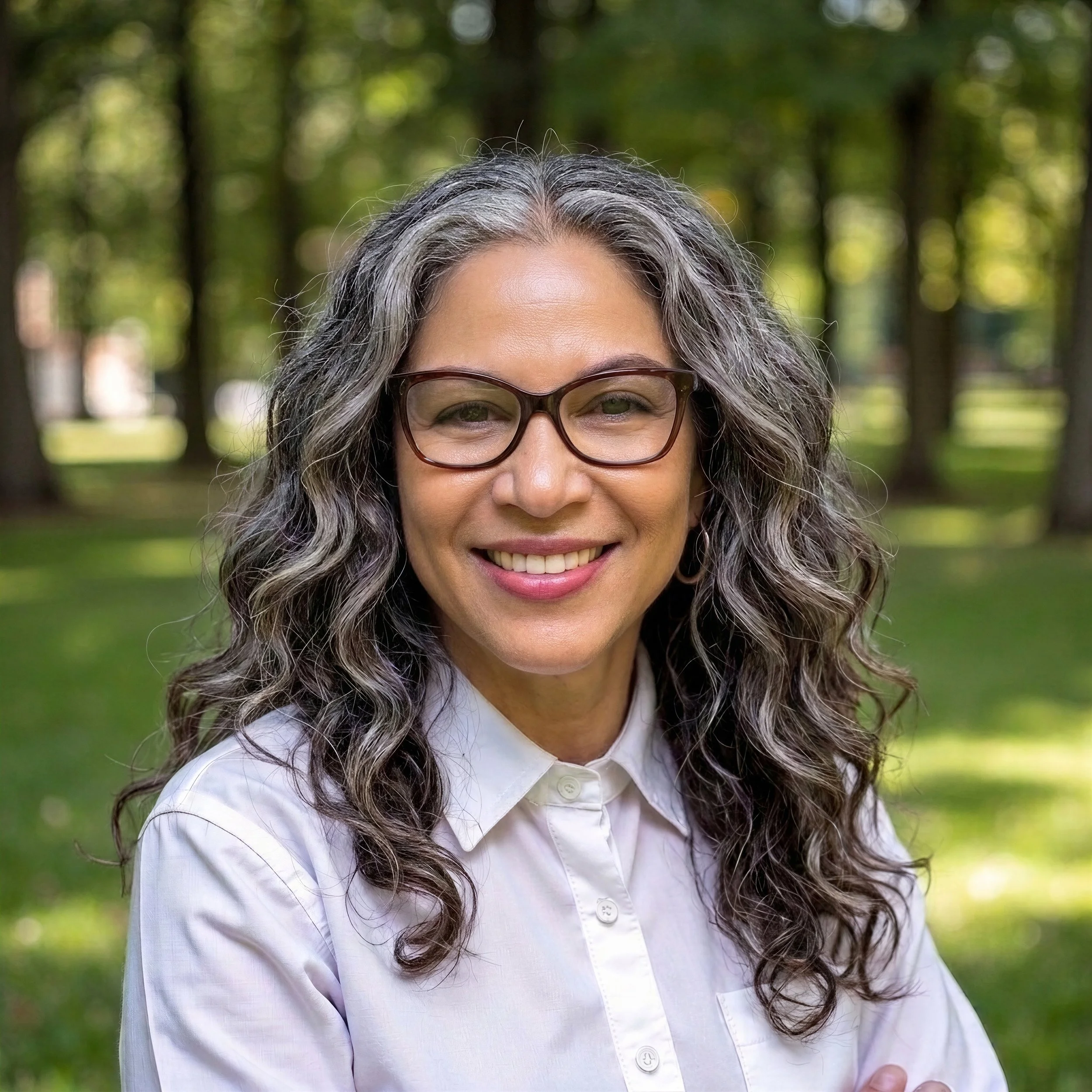 Close-up of an elderly woman with long gray hair, wearing glasses, dressed in a white shirt, smiling, outdoors with a blurred background of trees and greenery.