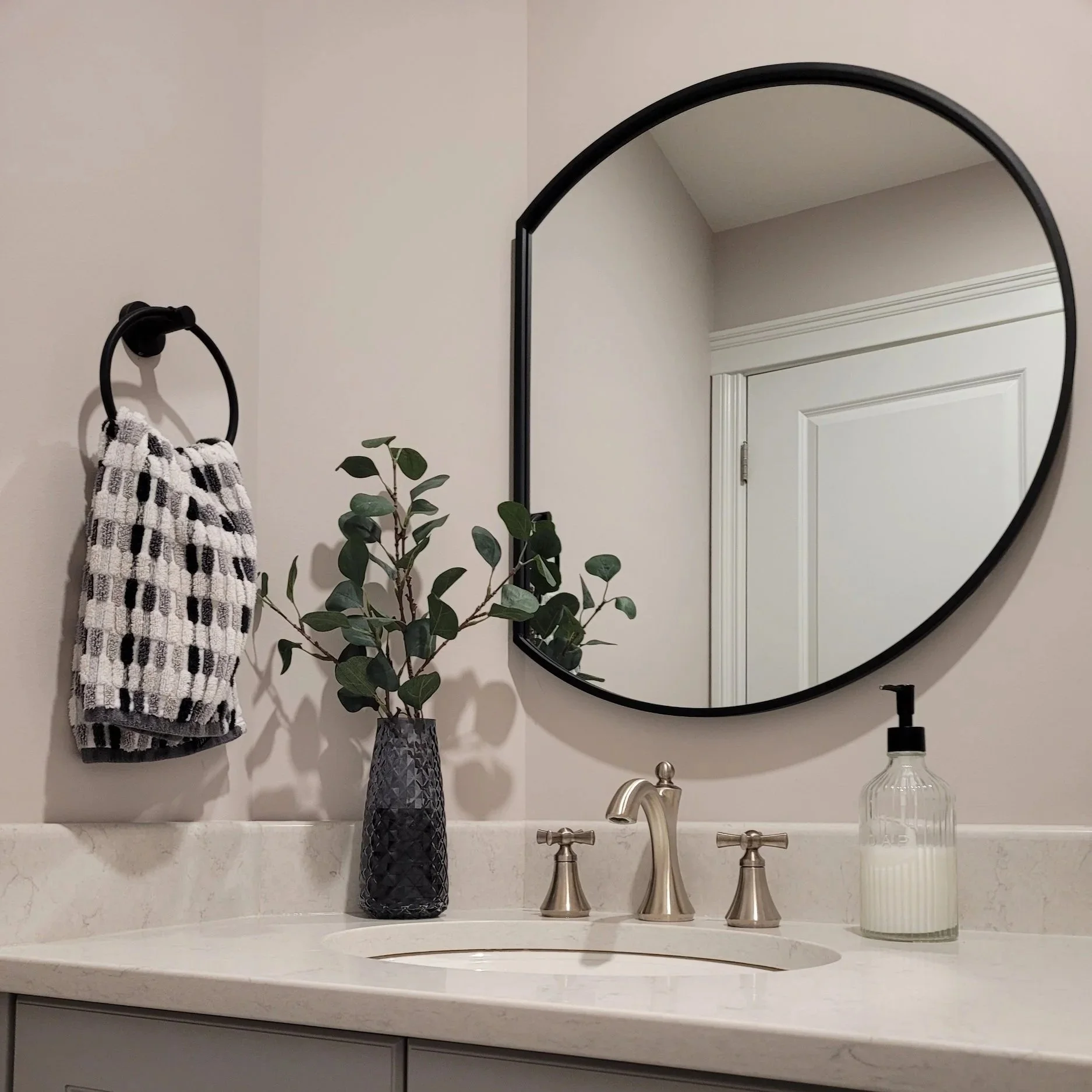 Bathroom sink vanity with a round mirror, a beige marble countertop, a tall textured black vase with a green leafy plant, a soap dispenser, and a small bottle. A towel hangs on a black ring, and the wall has a white door frame with beige walls.