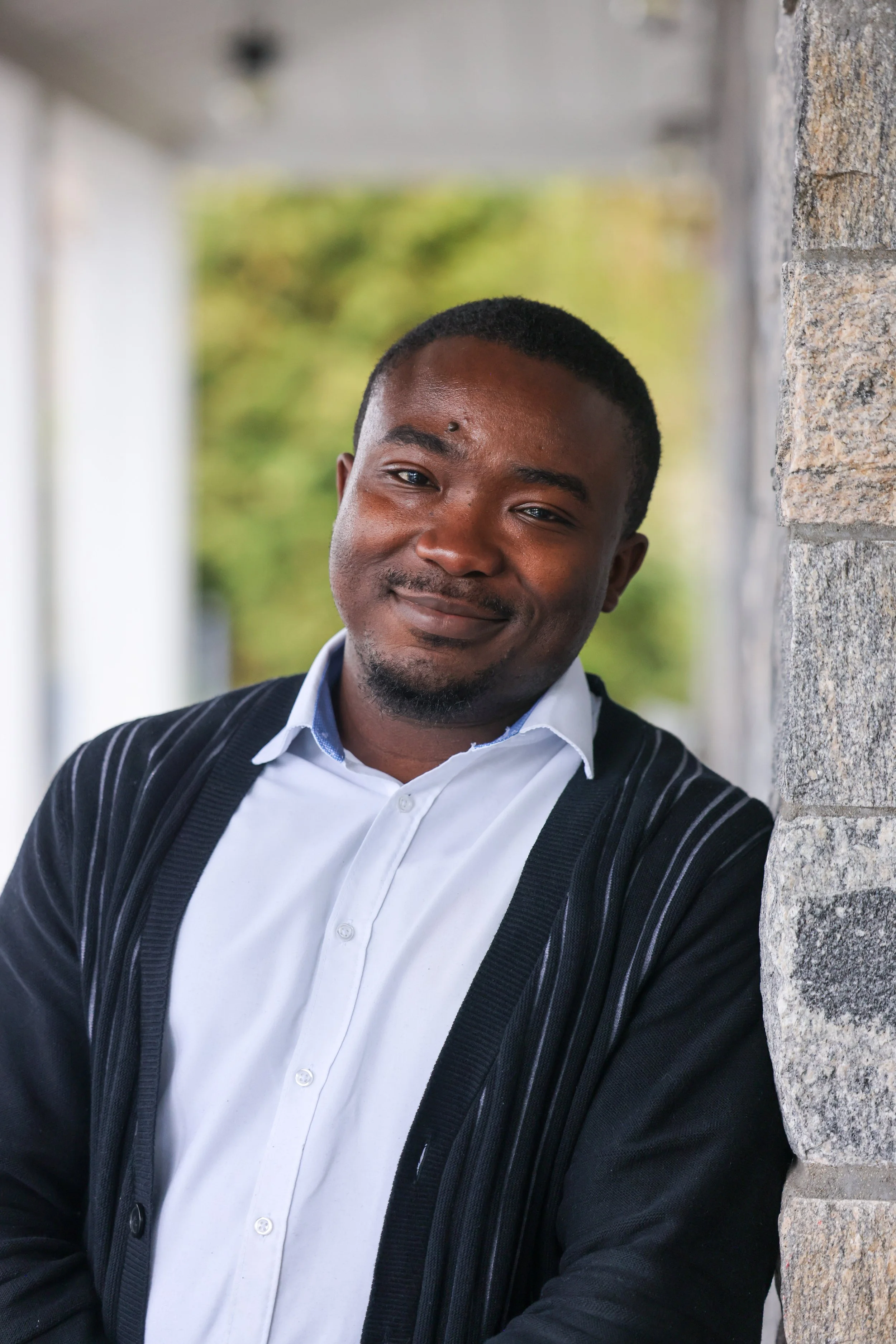 Portrait of a young man smiling, leaning against a stone wall with a blurred background of green trees.