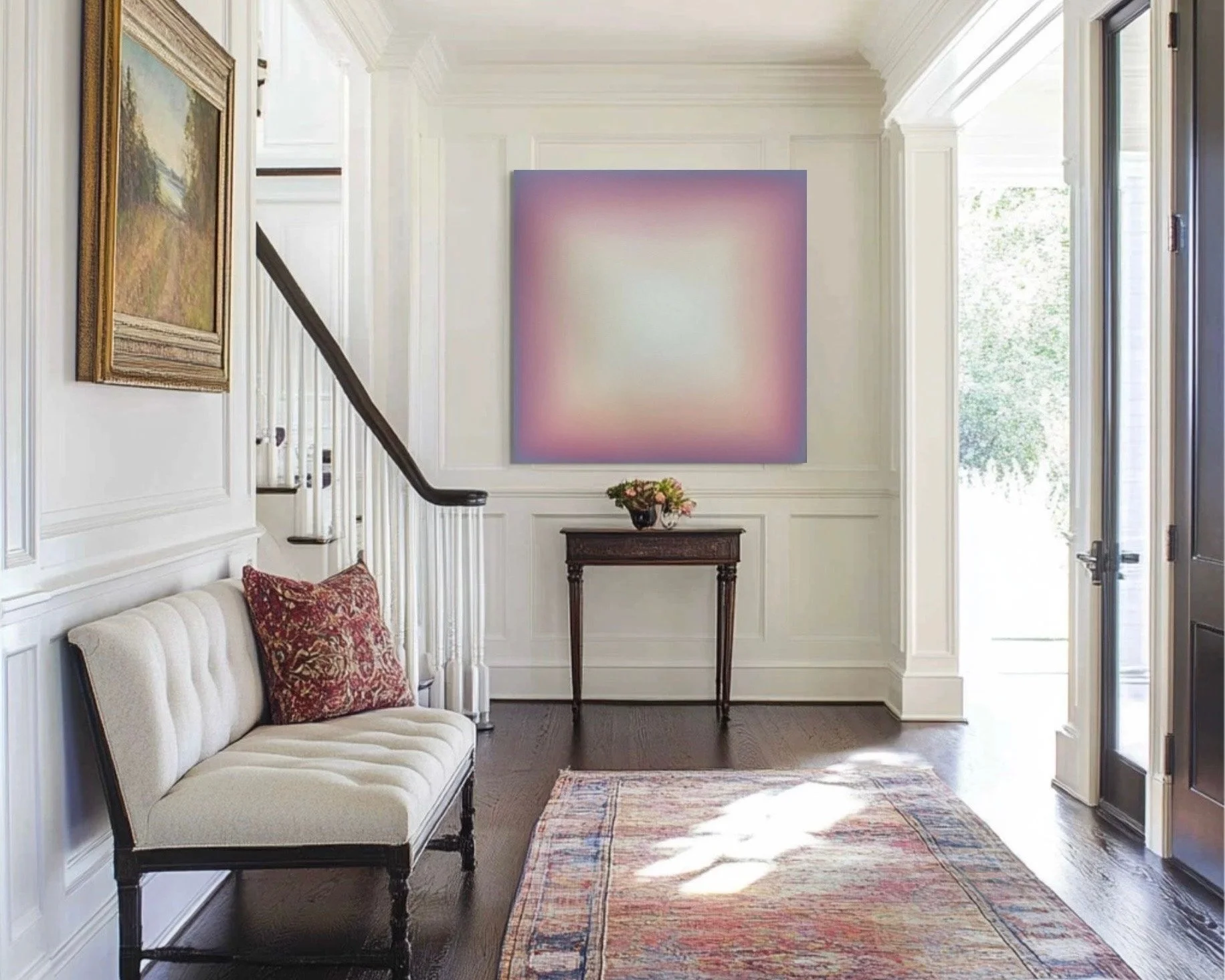 A well-lit entryway with cream-colored walls, dark hardwood flooring, and white trim. There's a small table with a flower arrangement, a cream sofa with red patterned pillows, a colorful rug, and framed artwork on the walls.