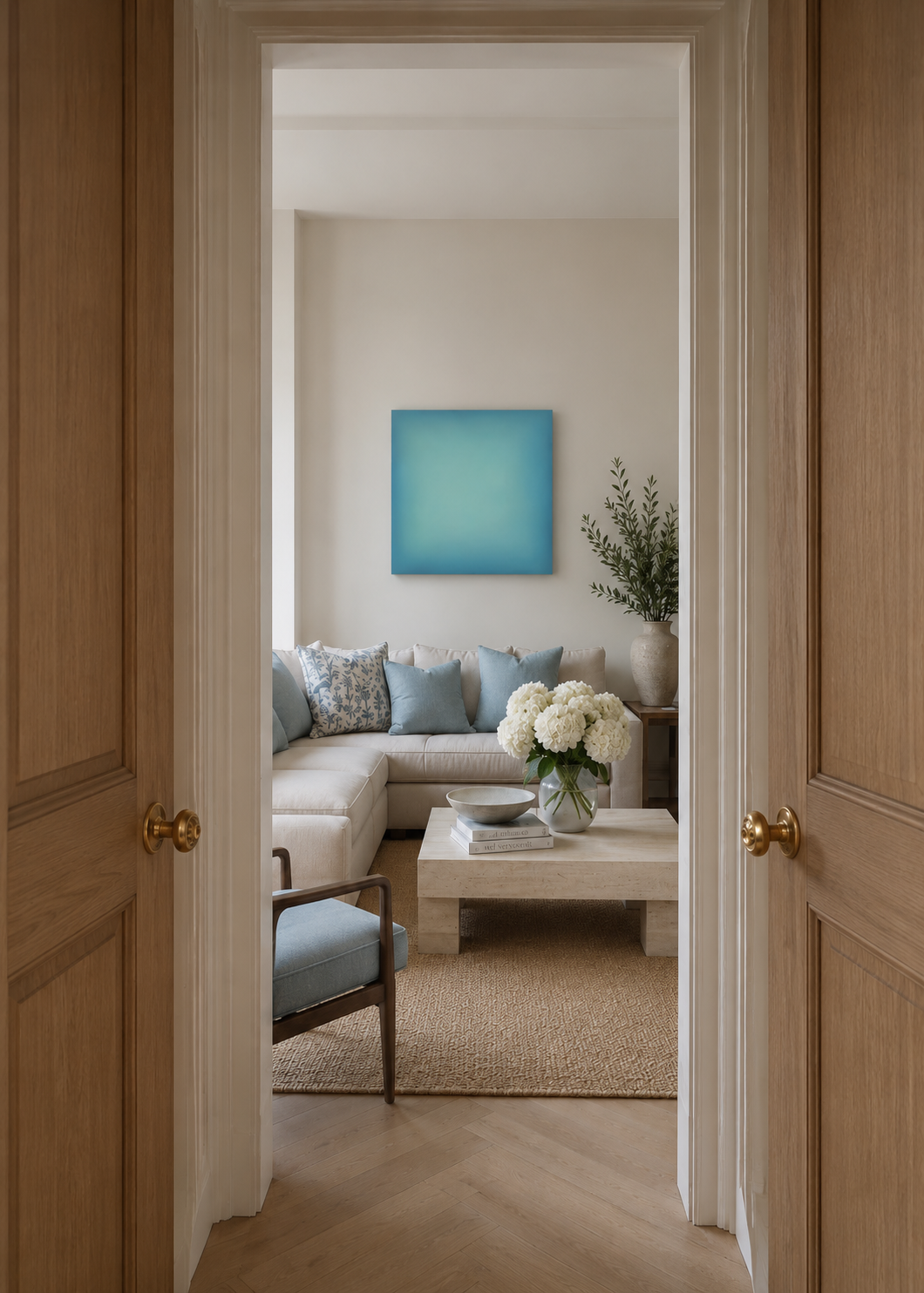 Living room with white sofa, blue and patterned cushions, a beige coffee table with white flowers, ceramic bowl, books, a large potted plant, a blue square wall art, and natural light.