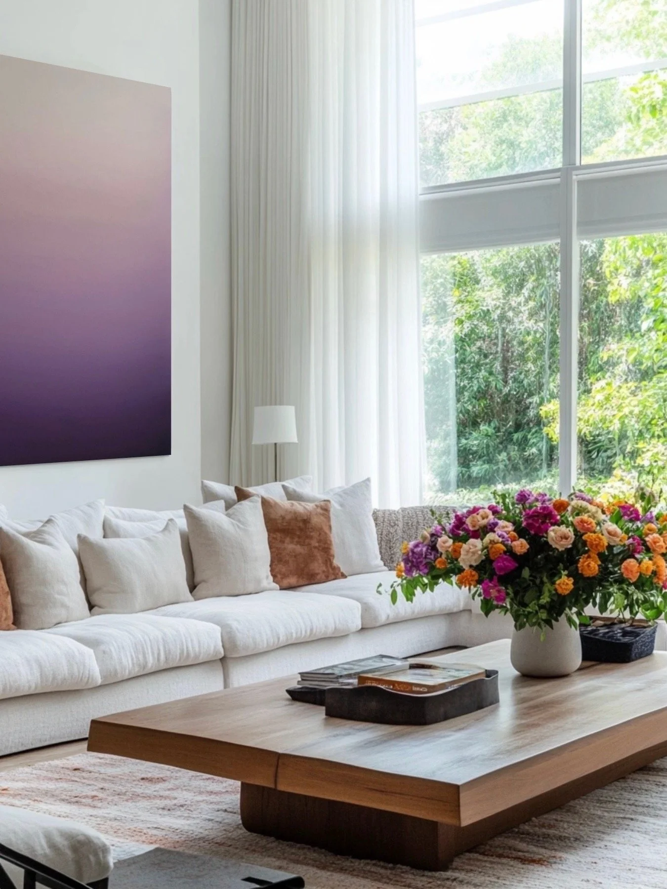 A living room with a white sofa, beige and pink pillows, a large window with sheer white curtains, a wooden coffee table with a black tray and magazines, and a large colorful flower arrangement on the table.
