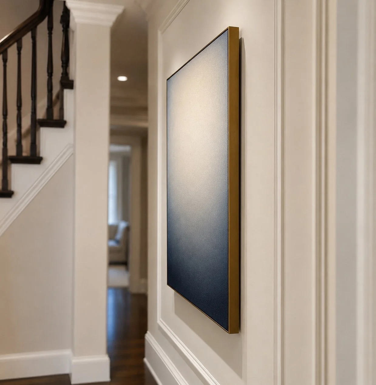 Close-up of a large blue canvas painting with a gold frame mounted on a white wall in a hallway, featuring a staircase and a hallway with a window and furniture in the background.
