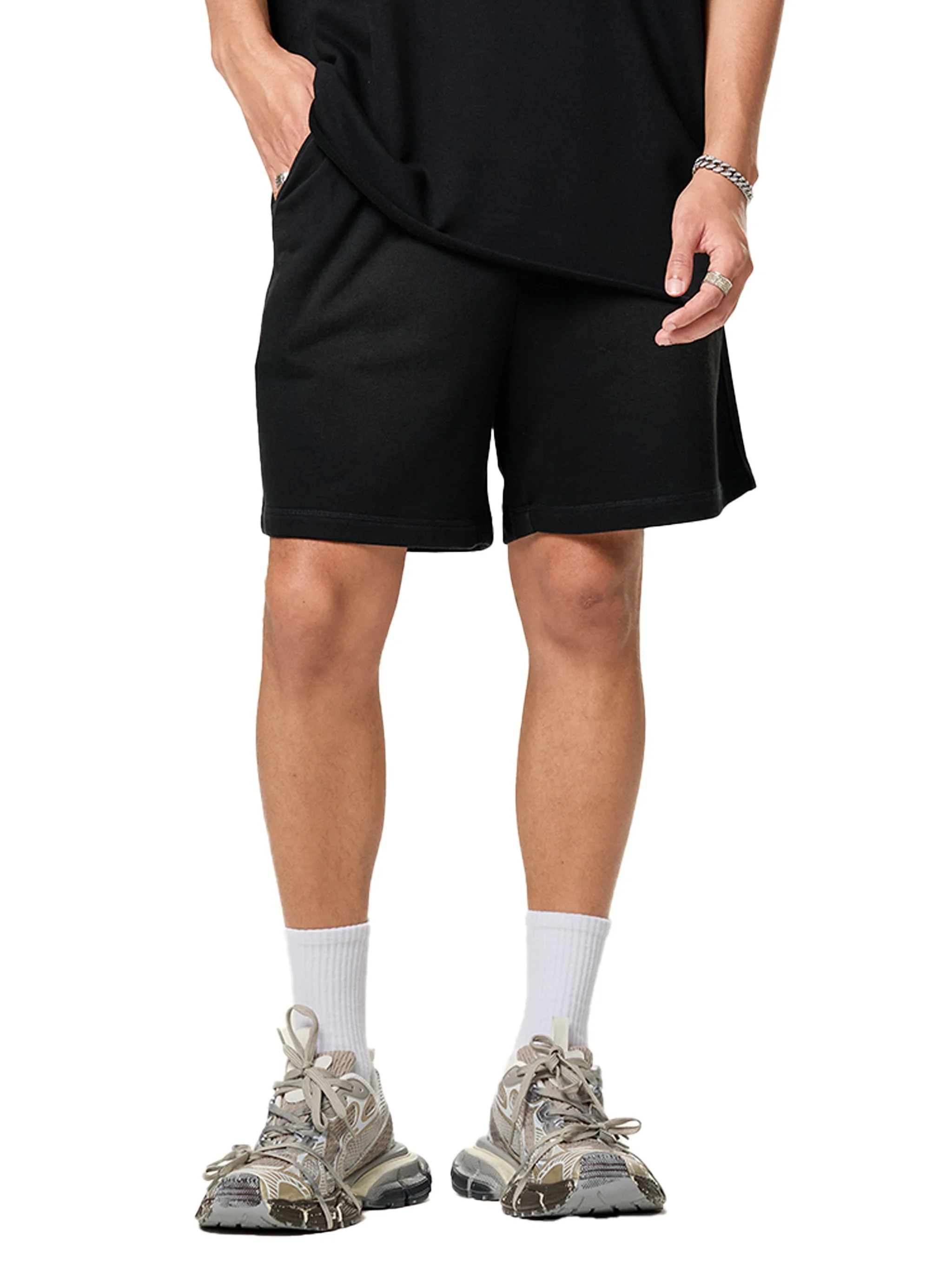 Person wearing beige and white sneakers, white crew socks, black shorts, and a black shirt, with tattoos on their legs and arms, and wearing rings and a bracelet.
