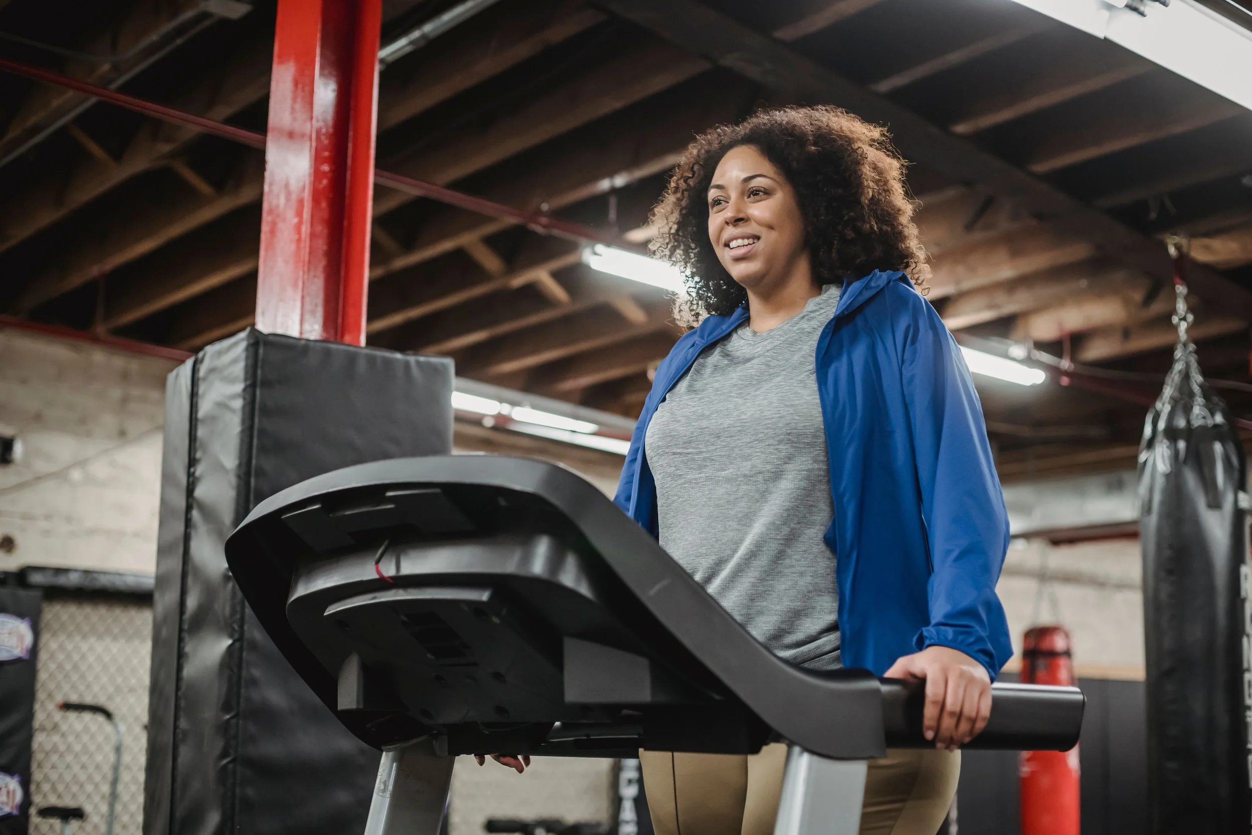 Woman in a blue jacket and gray T-shirt standing beside a treadmill in a gym.