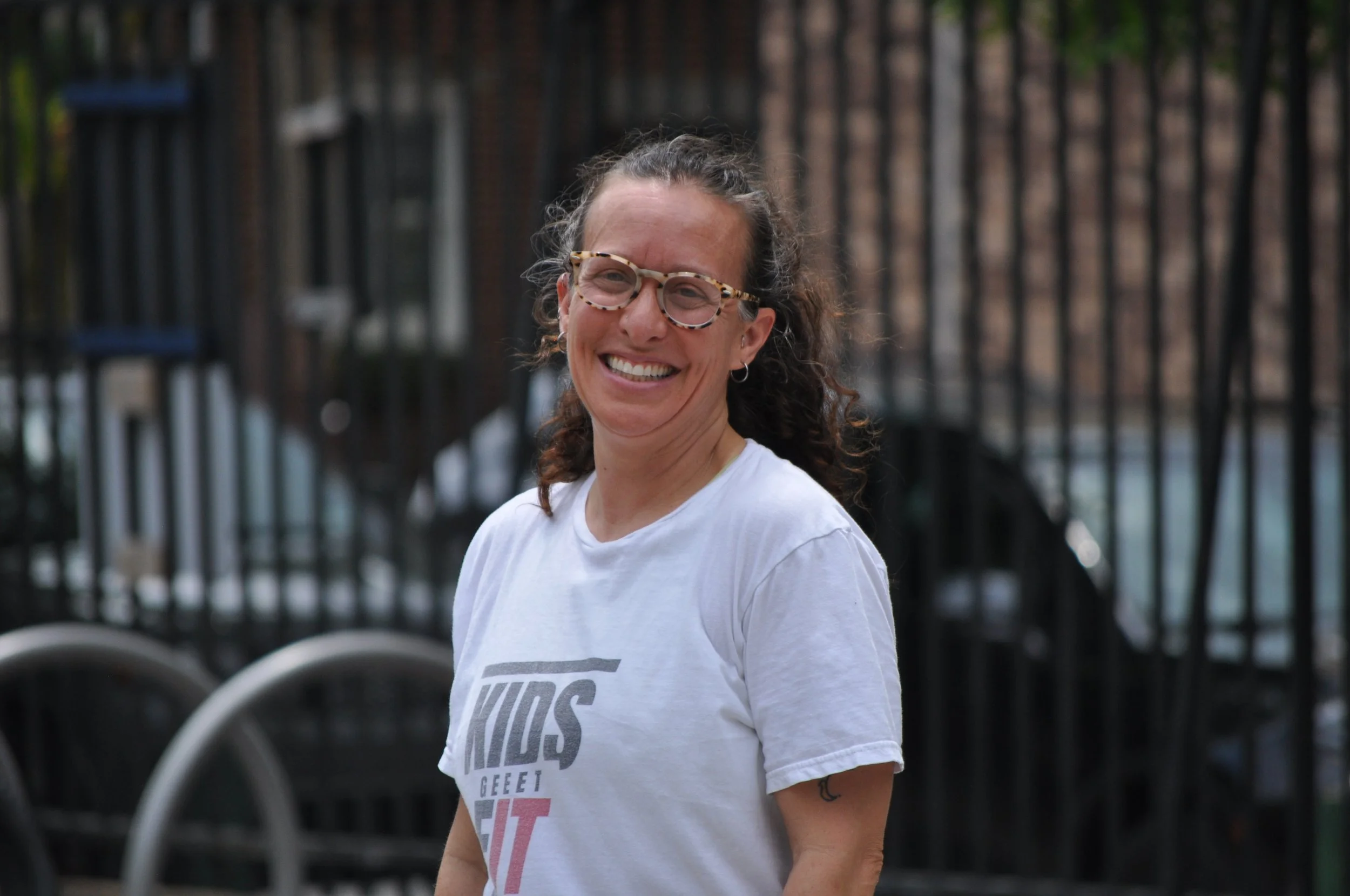 A smiling woman with curly hair and glasses wearing a white T-shirt standing outdoors in front of a black fence and blurred background.