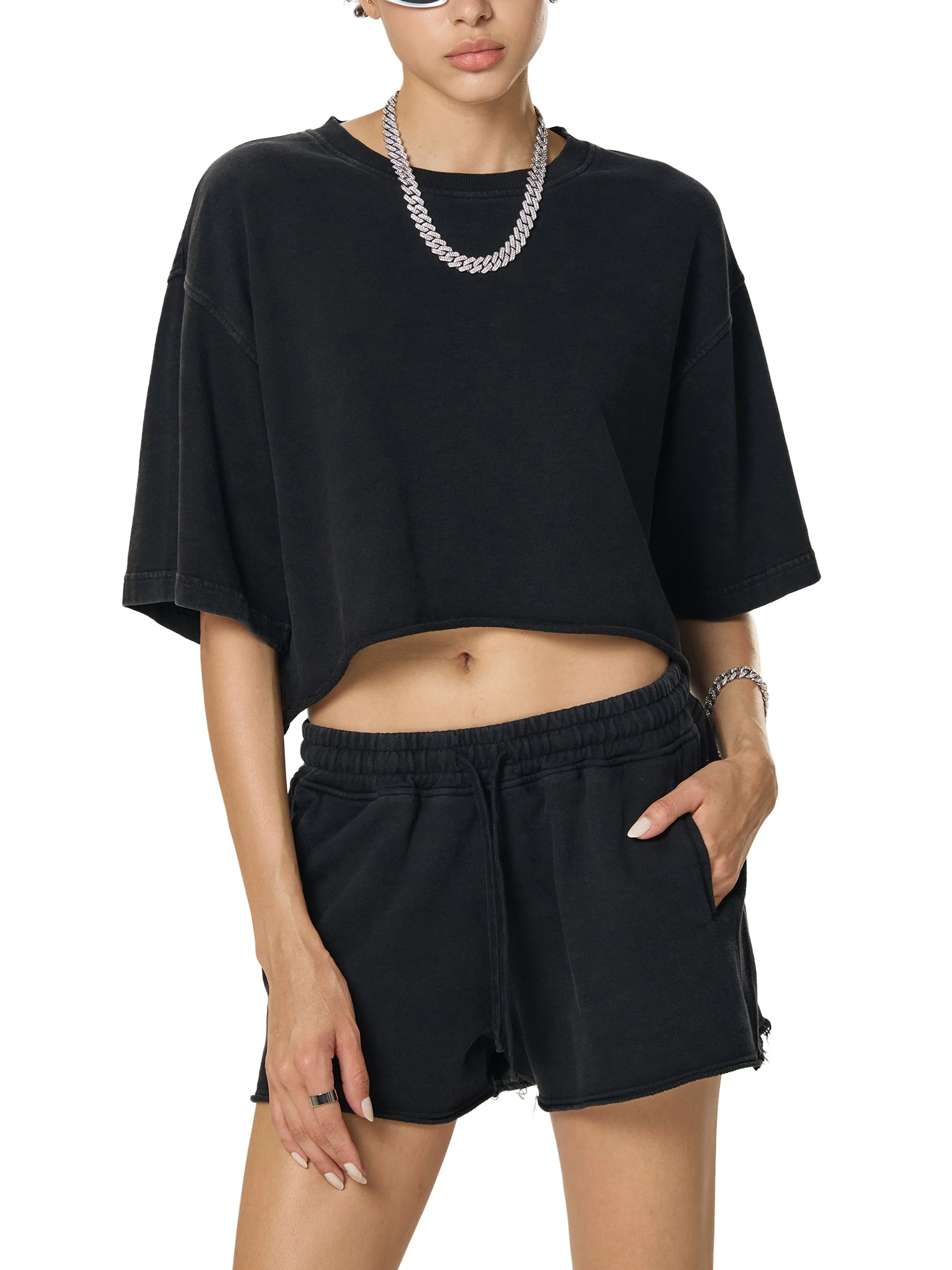 A woman wearing a black cropped t-shirt, black shorts, white sunglasses, a silver chain necklace, and a silver bracelet.