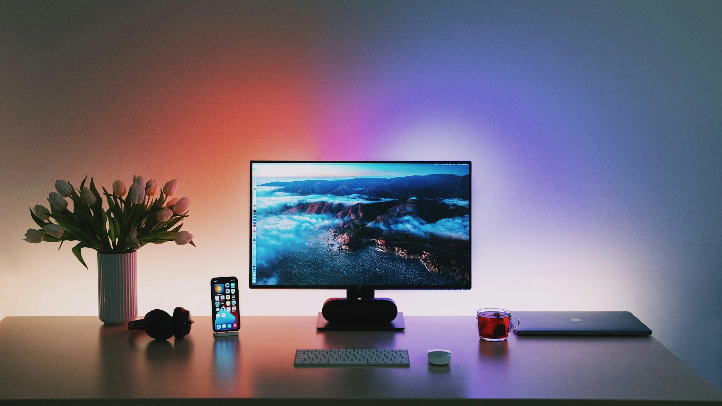A modern desk setup featuring a large monitor displaying landscape scenery, a smartphone with app icons, a pair of black headphones, a white keyboard, a white mouse, a silver laptop, a glass mug with red beverage, and a vase with pink tulips, against a wall illuminated by colorful ambient lighting.