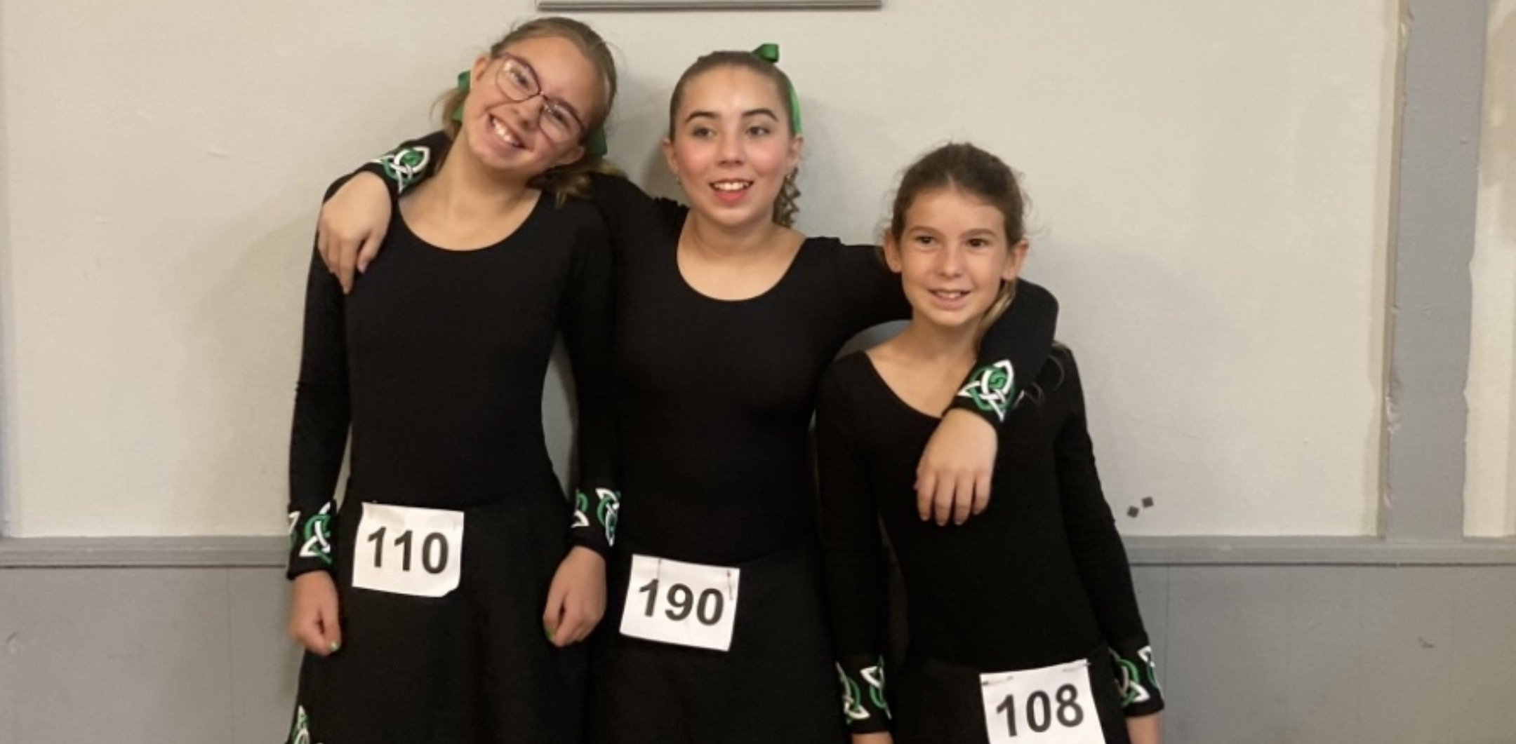 Three young girls in black long-sleeve shirts and black skirts standing together with arms around each other, smiling. They are wearing signed numbers 110, 190, and 108 on their skirts and have green hair bows and accessories with a green, white, and black pattern. They appear to be at a dance or cheerleading event indoors against a plain wall.