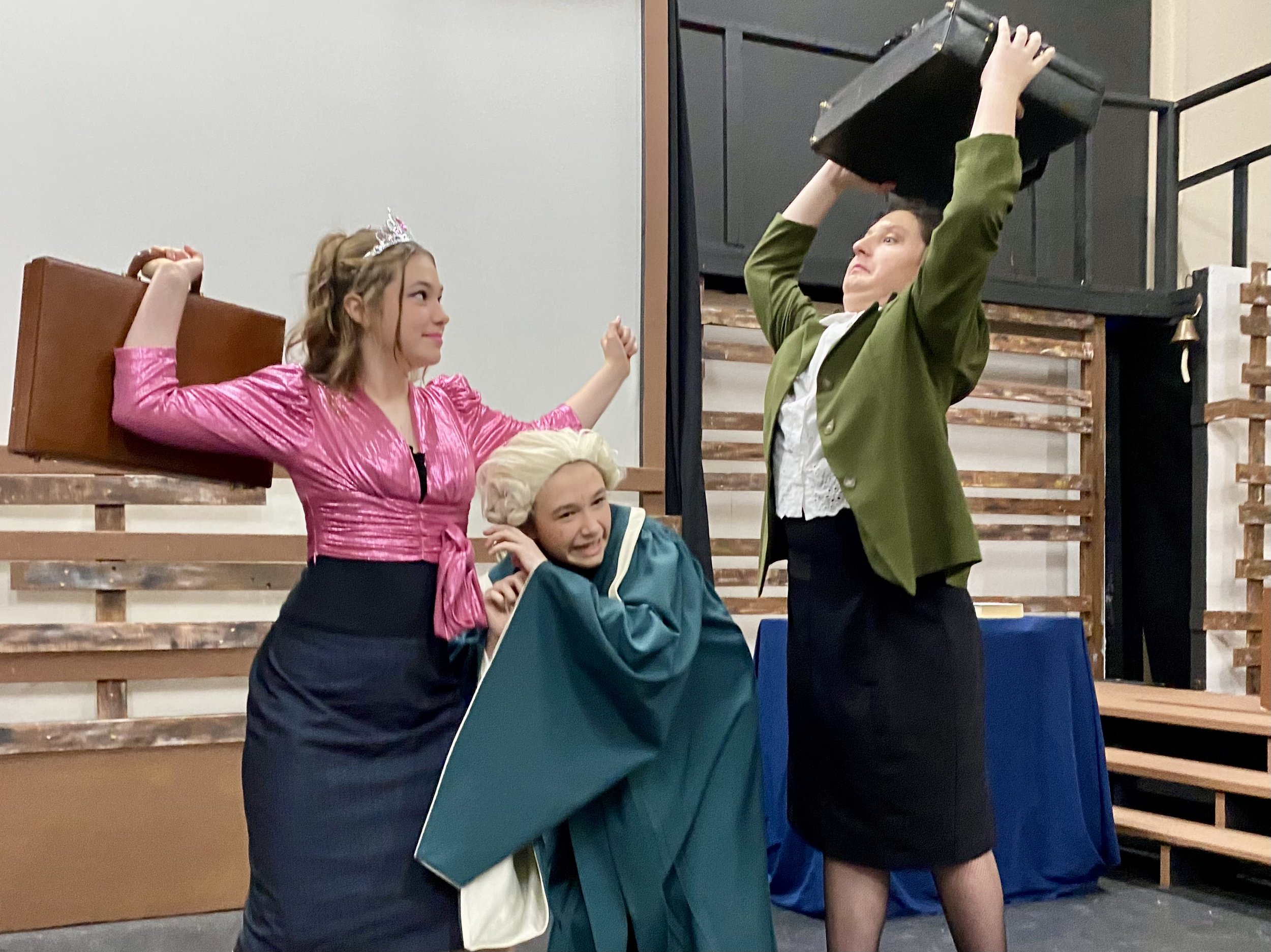 A young girl dressed as a princess with a tiara, purple dress, and pink sash, holding a small brown suitcase, is smiling. An older woman in a green jacket and white shirt is raising a black suitcase above her head, appearing to be in a playful or theatrical pose. There are other children in the background, and the setting appears to be a stage or a decorated environment with wooden structures.