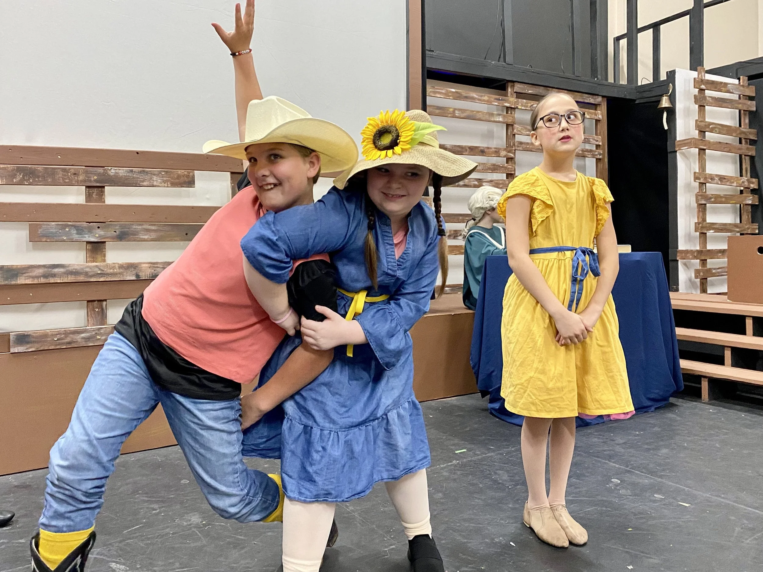 Three children in colorful costumes, two girls in large straw hats with sunflower decorations, and a girl in a yellow dress, standing in a theater or performance space with wooden seating and stage decorations.