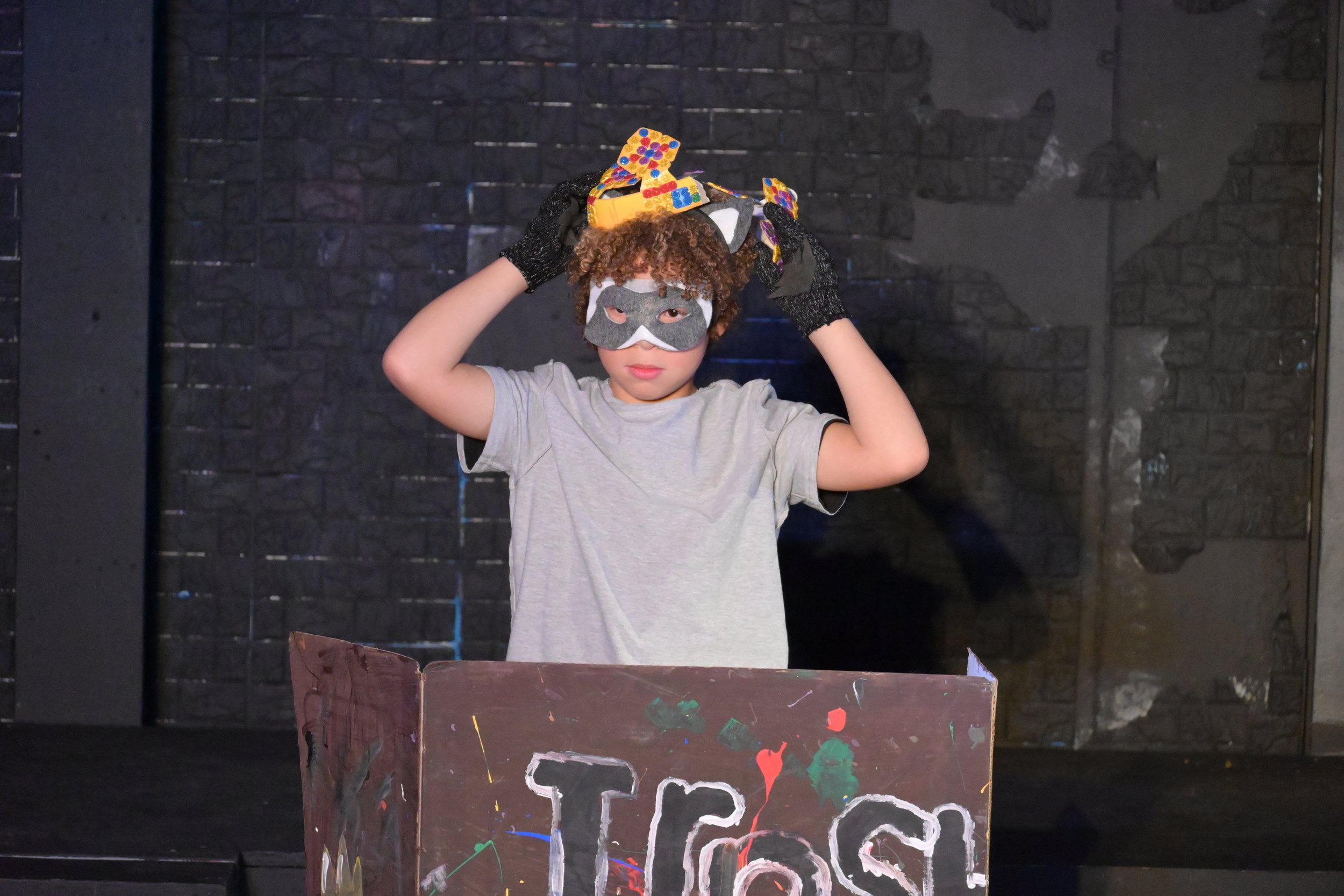 A child wearing a galaxy-themed mask and black gloves puts a paper crown with colorful dots on their head, standing behind a table covered with colorful paints and art supplies, in front of a dark brick wall.