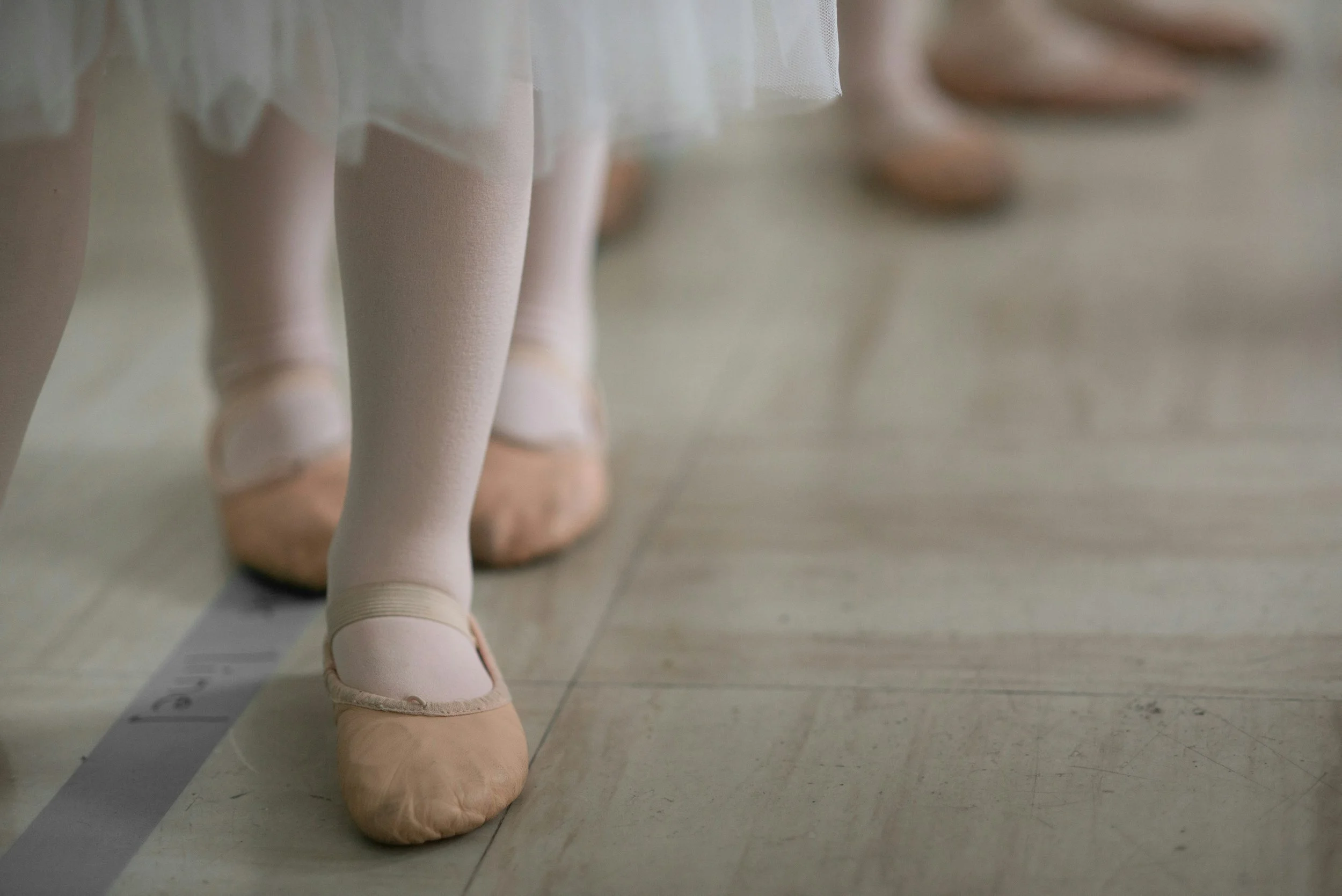 Close-up of ballet dancer's feet in pink slippers, standing on tiptoes on a tiled dance studio floor, with a measuring tape visible in the background.