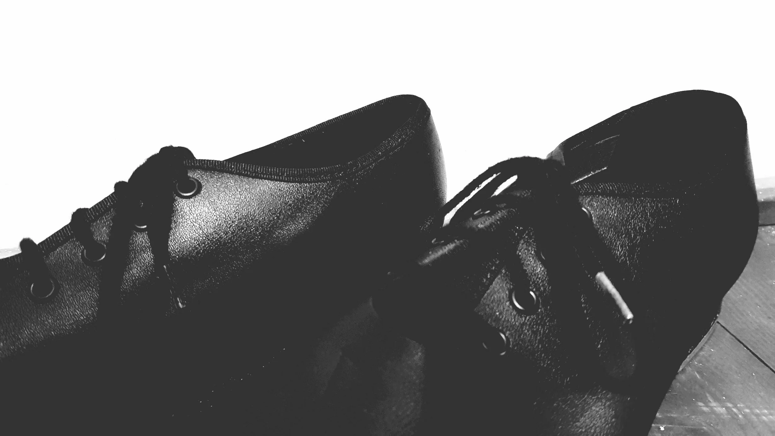 Close-up of a pair of black leather combat boots on a wooden floor, in black and white.