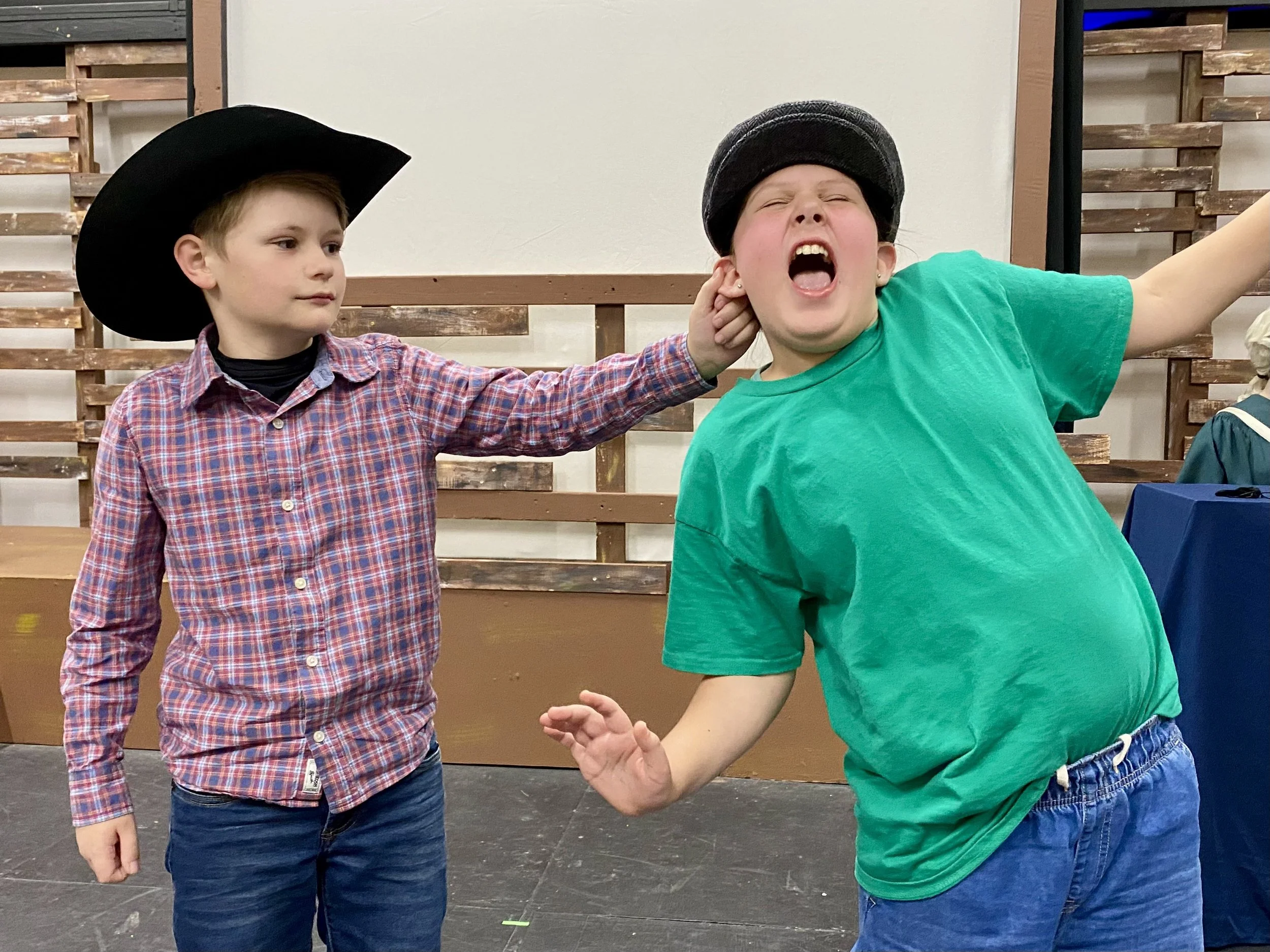 Two young boys playing together, one wearing a black cowboy hat and plaid shirt, the other wearing a gray cap and bright green T-shirt. The boy in the green shirt is shouting or singing with his mouth open wide. They are indoors with wooden paneling in the background.