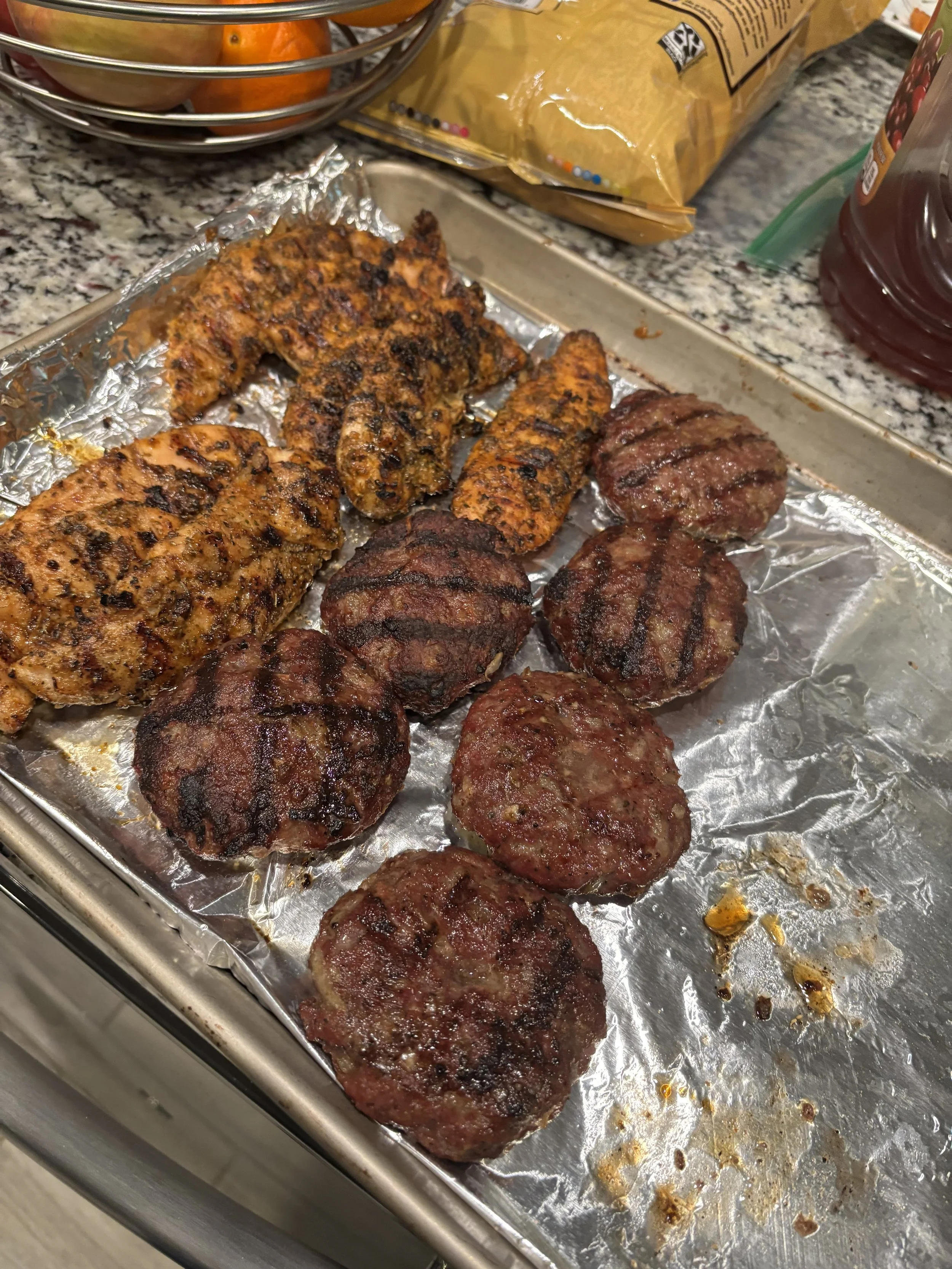Cooked meat patties and grilled chicken on a baking sheet lined with aluminum foil.