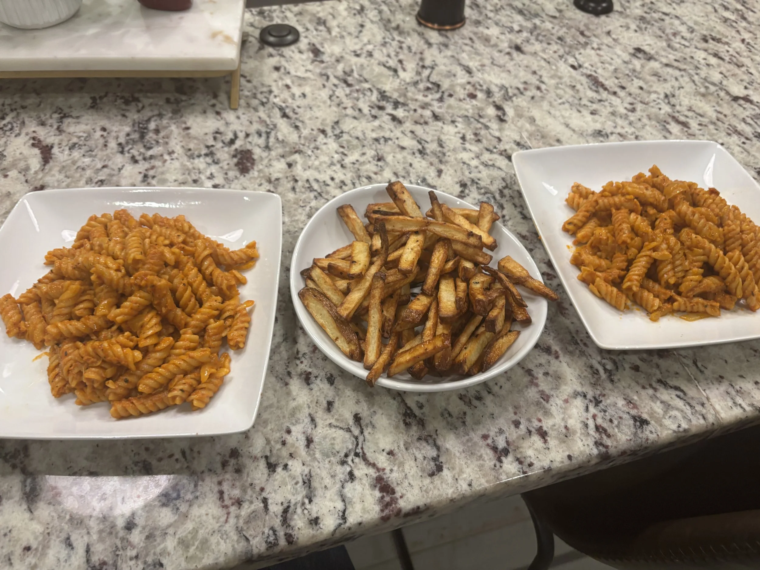 Three white plates on a granite countertop with pasta and French fries, and a white bowl of pasta in the middle