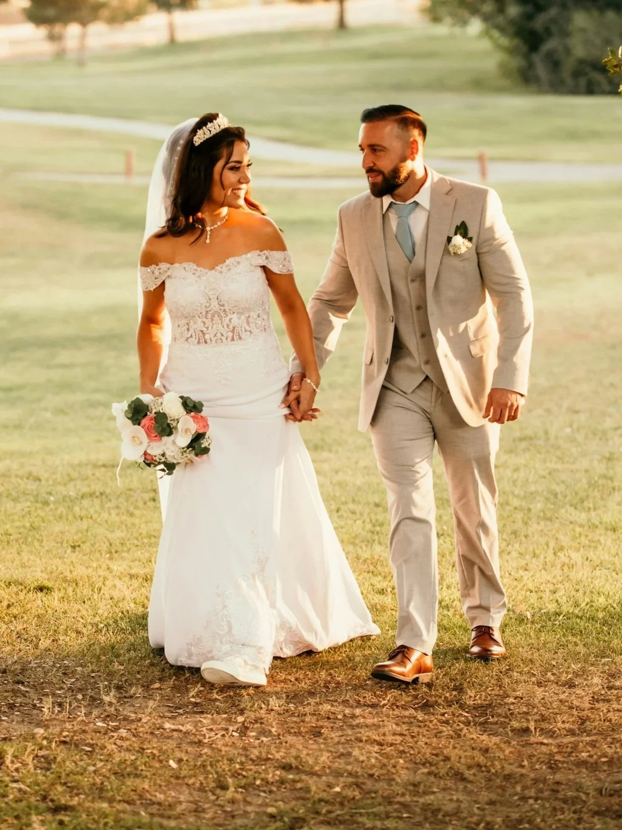 A bride and groom walk hand in hand outdoors during sunset. The bride wears a white lace off-shoulder wedding dress and holds a bouquet, while the groom wears a light beige suit and brown shoes.