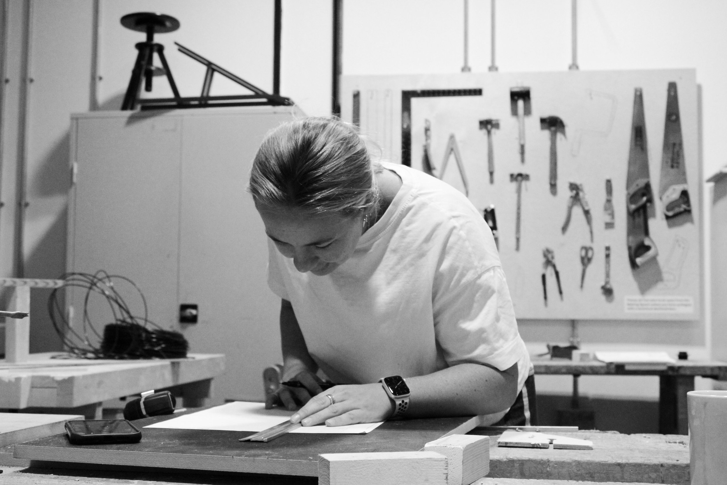 Person working at a workshop table with tools on a pegboard behind them in black and white.