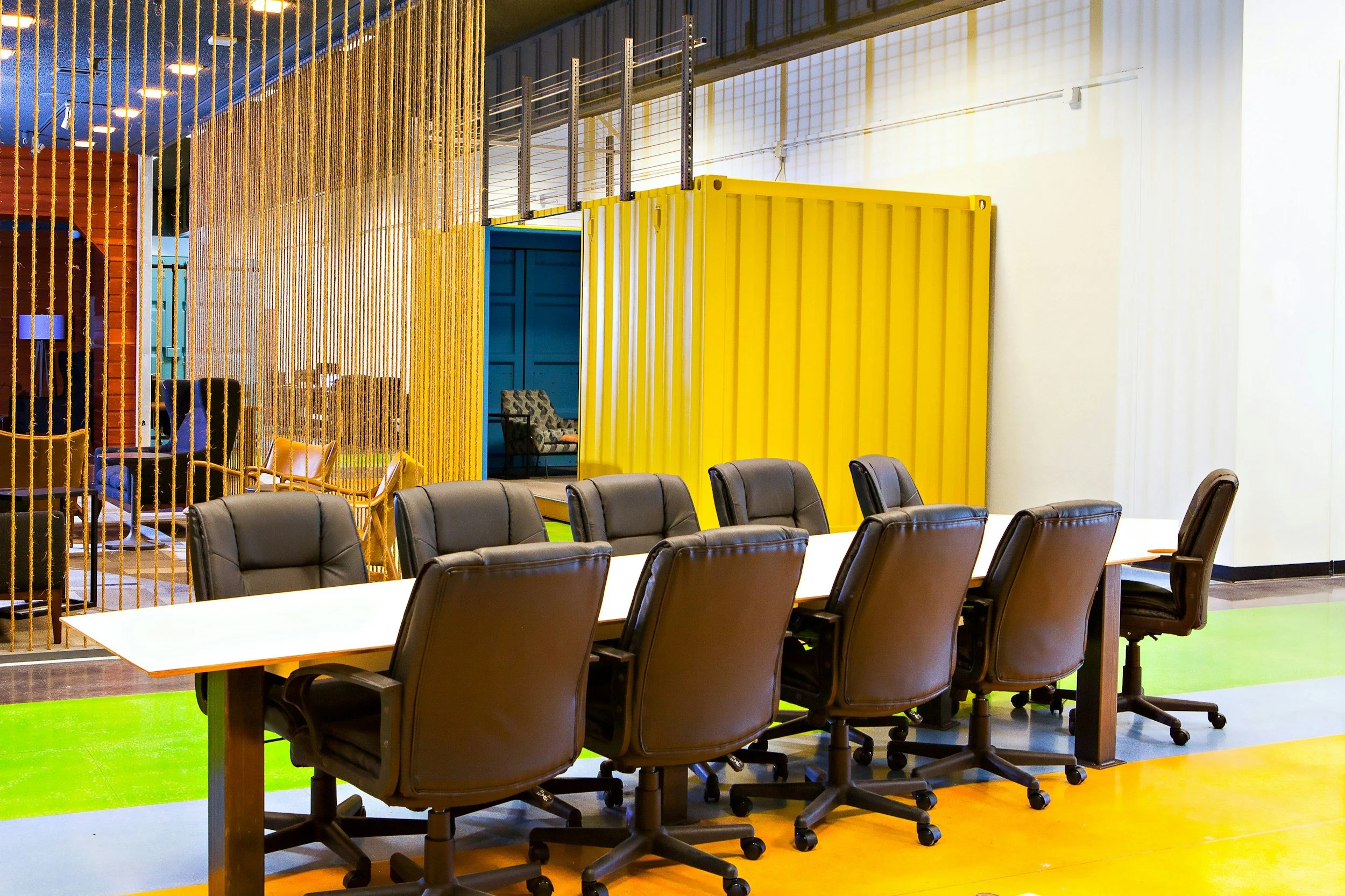 Conference room with black chairs around a long white table, colorful flooring, yellow and blue walls, and decorative hanging yellow strings.