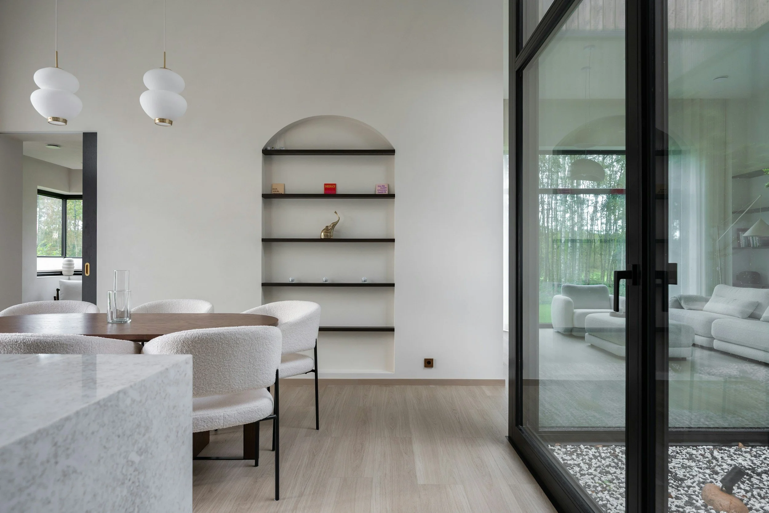 Modern living room with white walls, two hanging pendant lights, a built-in shelf with small decor items, and large glass sliding doors leading to an outdoor patio with white furniture.