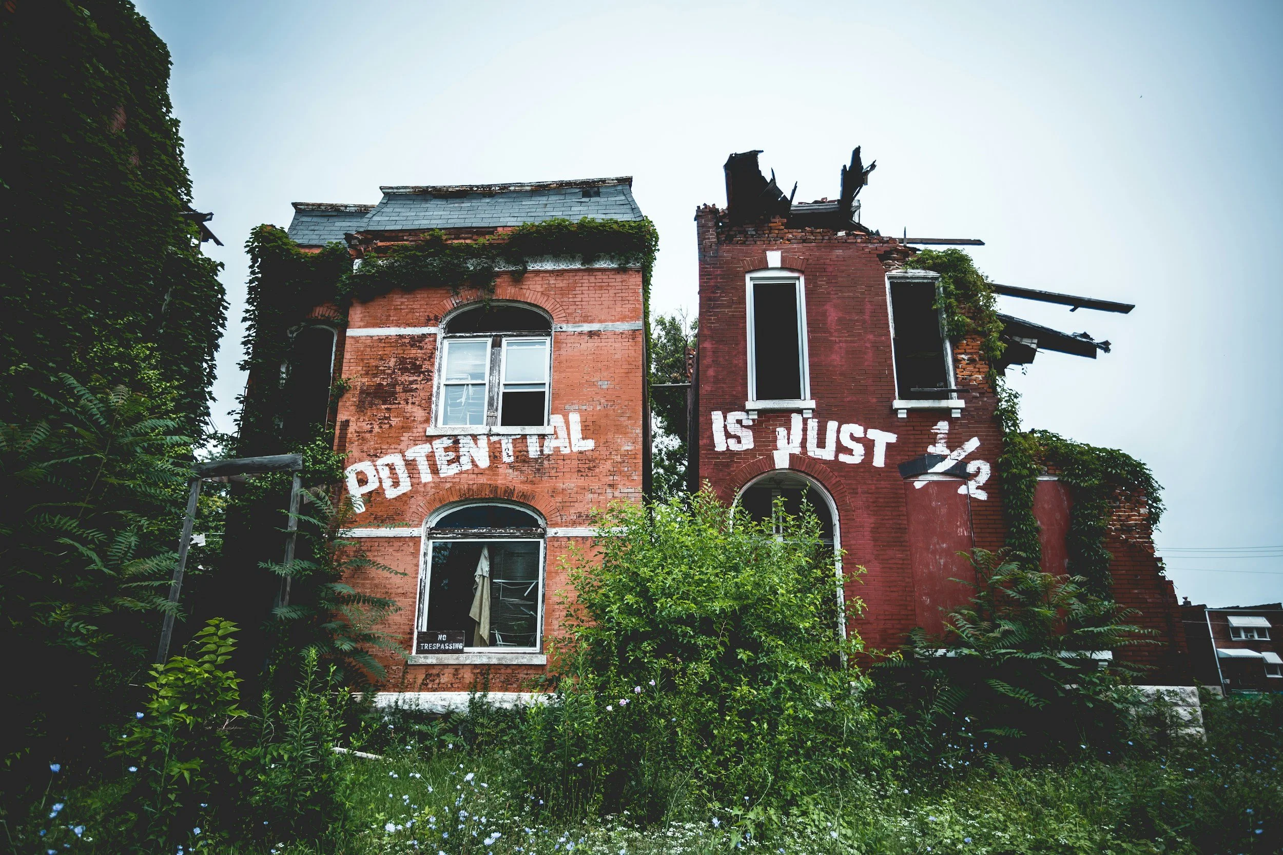 Two old brick buildings with broken and missing roof sections, overgrown with vegetation, with faded white painted signs on the front reading 'POTENTIAL IS JUST 1/2'.