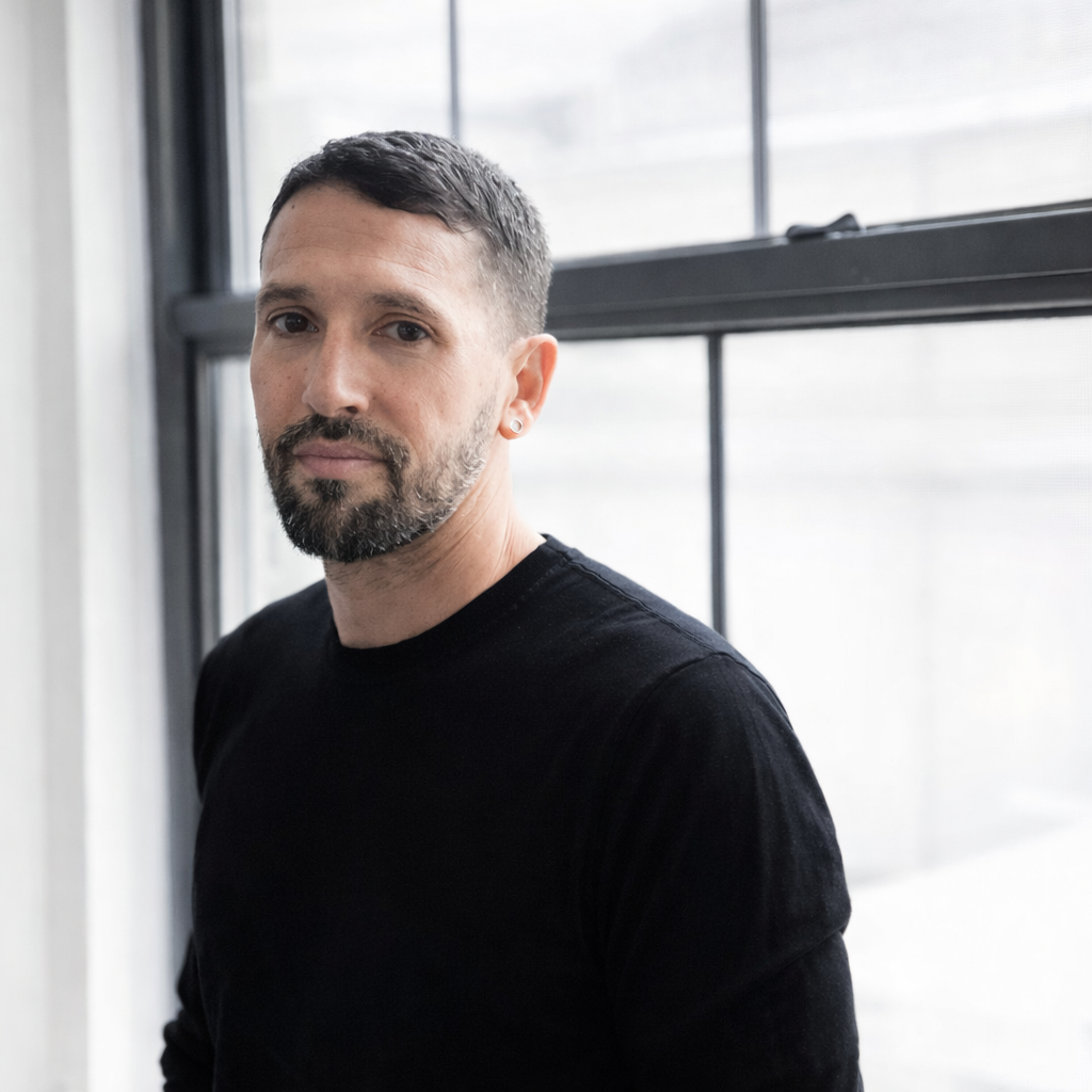 A man with short dark hair and a beard, wearing a black shirt, standing indoors near a window with blinds.