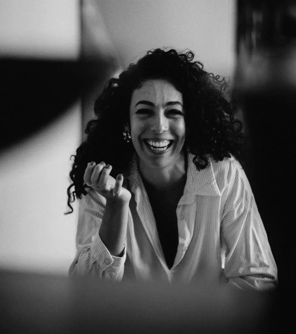 Black and white photo of a woman with curly hair smiling and pointing towards the camera, wearing a striped shirt.
