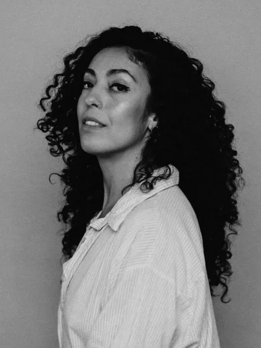 A woman with curly hair wearing a light-colored shirt, looking at the camera with a confident expression.