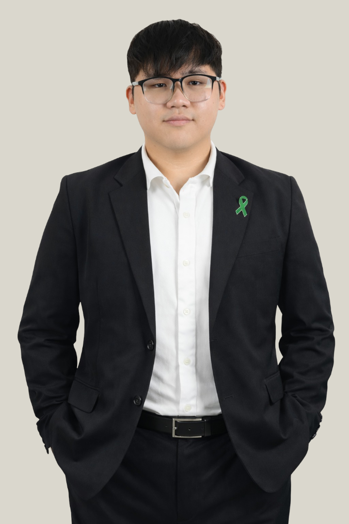 A young man wearing glasses, a black suit, white shirt, with hands in pockets, standing against a plain background, with a green awareness ribbon pin on his lapel.