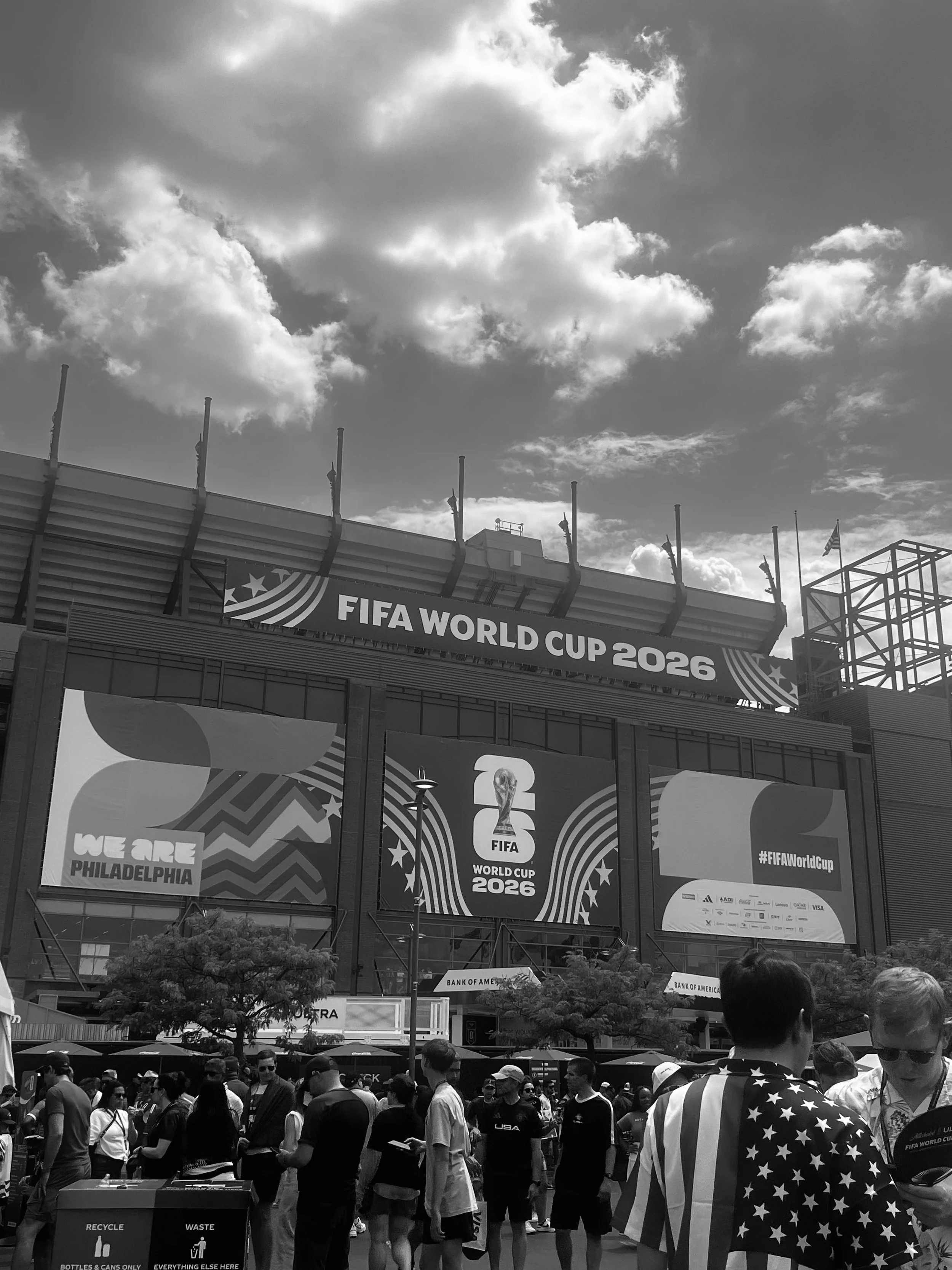 Crowd of people gathered outside a football stadium with FIFA World Cup 2026 banners and signs, some wearing American flags.