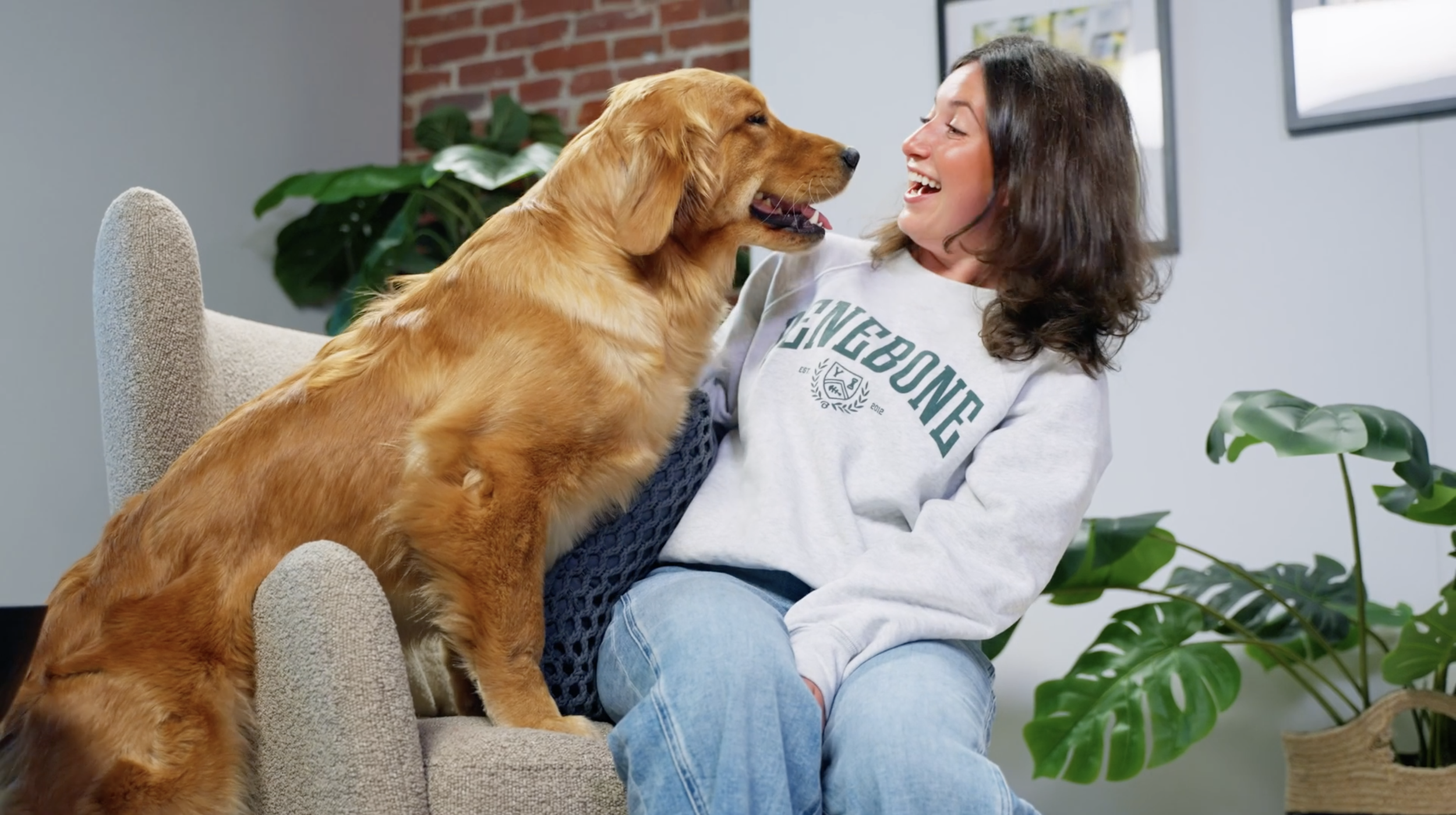 A woman with dark hair smiling and laughing while holding a golden retriever dog that is standing on a beige couch.