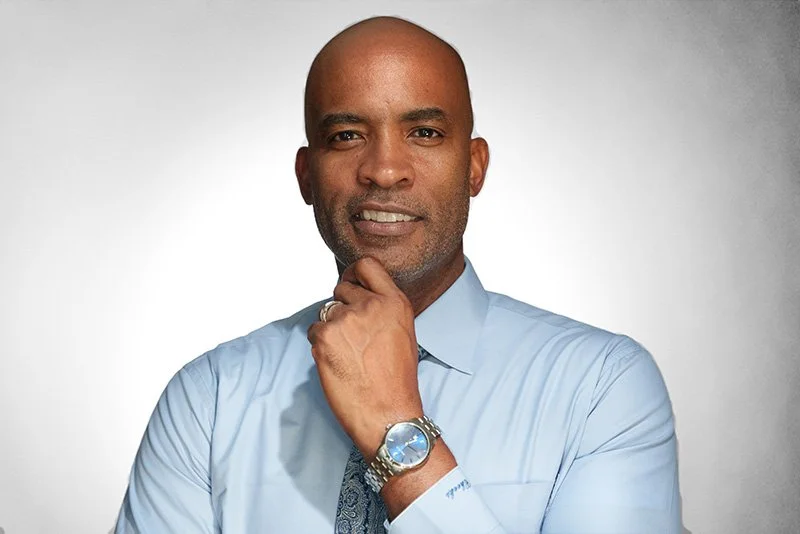 A confident African American man with a shaved head and beard, wearing a light blue dress shirt, a blue patterned tie, and a silver wristwatch, standing against a plain gray background.