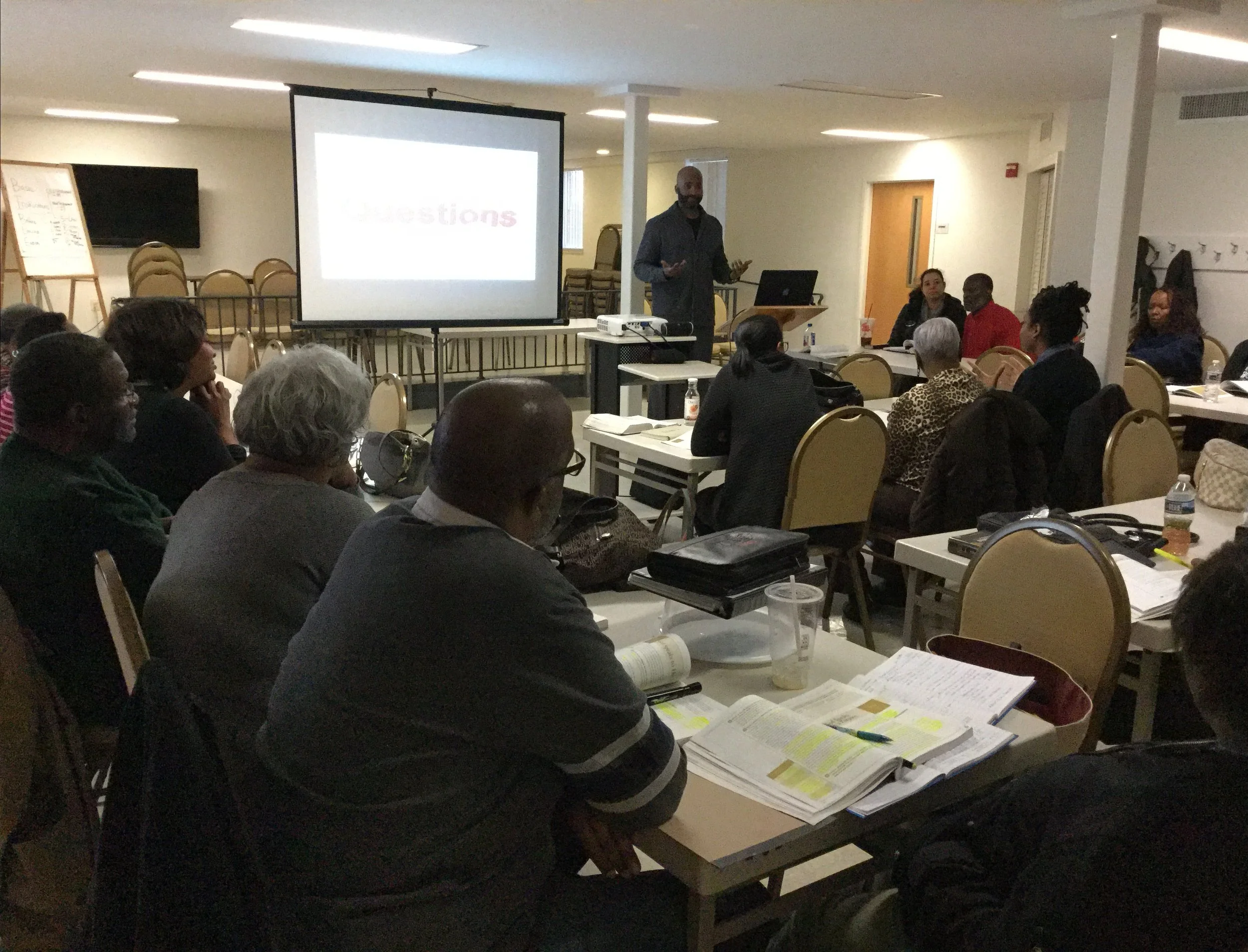 A man is giving a presentation to a group seated at tables in a conference room. The room has a large projector screen, chairs, and tables with documents and bottles of water.