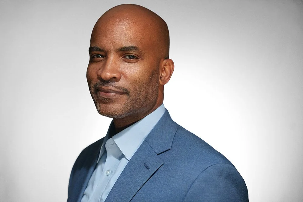 A professionally dressed man in a blue suit and light blue shirt, looking at the camera with a neutral expression, against a plain gray background.