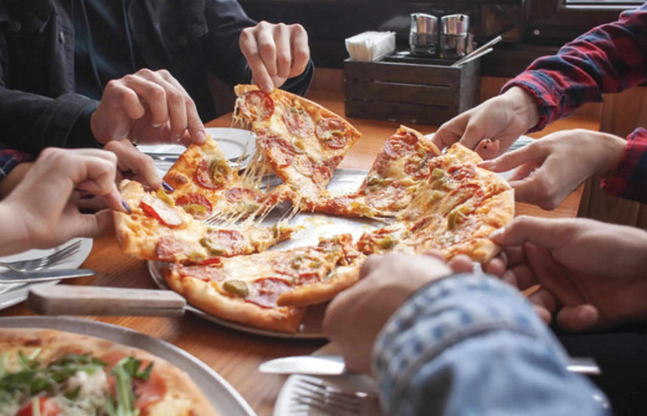 Several people are sharing a pizza in a restaurant. The pizza has pepperoni, green peppers, and melted cheese. Some are pulling slices while others are reaching for pieces. The setting is cozy with a wooden table and condiment containers in the background.