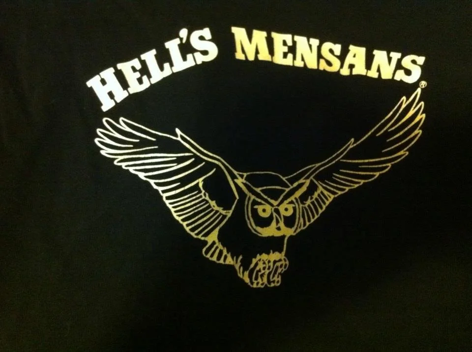 Black shirt with a yellow outline of an owl with spread wings, and the text "HELLO'S MENSAANS" above the owl in yellow and white letters.