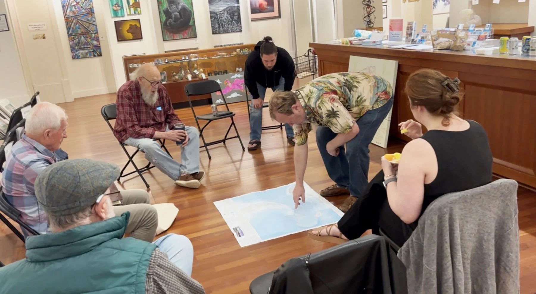 Group of people gathered in a room, sitting on chairs and engaging in a discussion or activity around a large map on the floor. The room has wooden flooring and colorful artwork on the walls, with display cases and informational materials in the background.