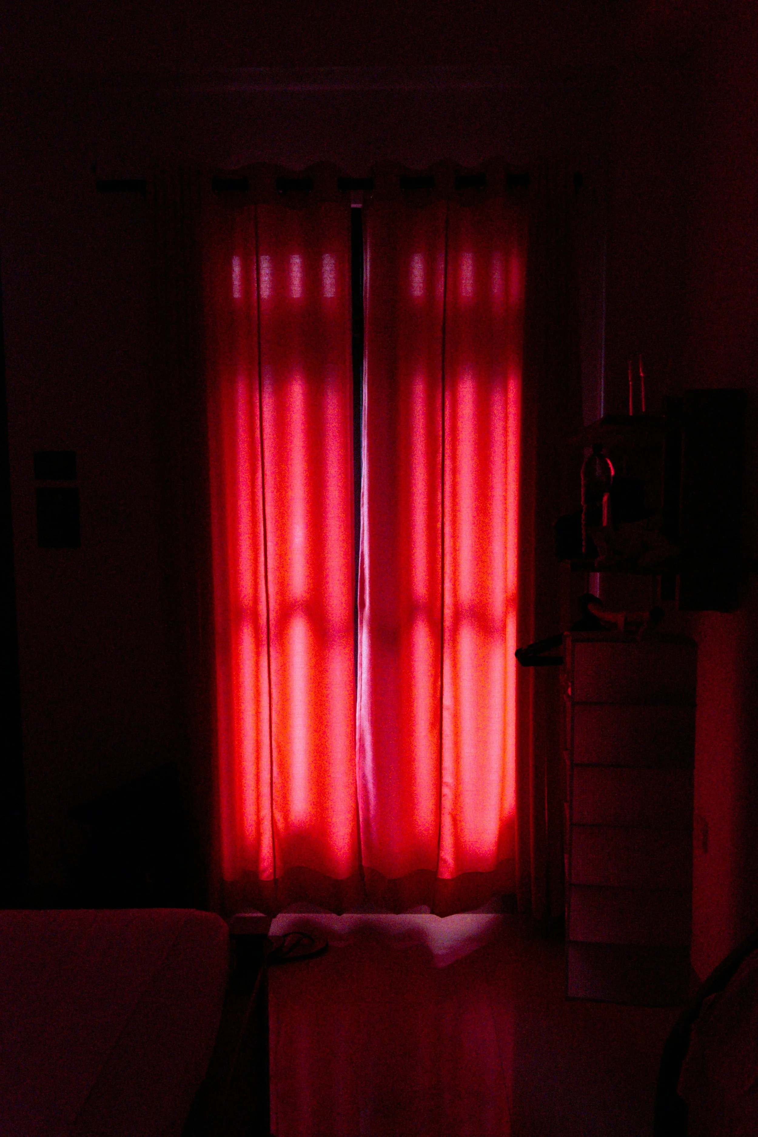 Red curtains in a dimly lit room with minimal light coming through, with furniture partially visible and shadows creating a dark ambiance.