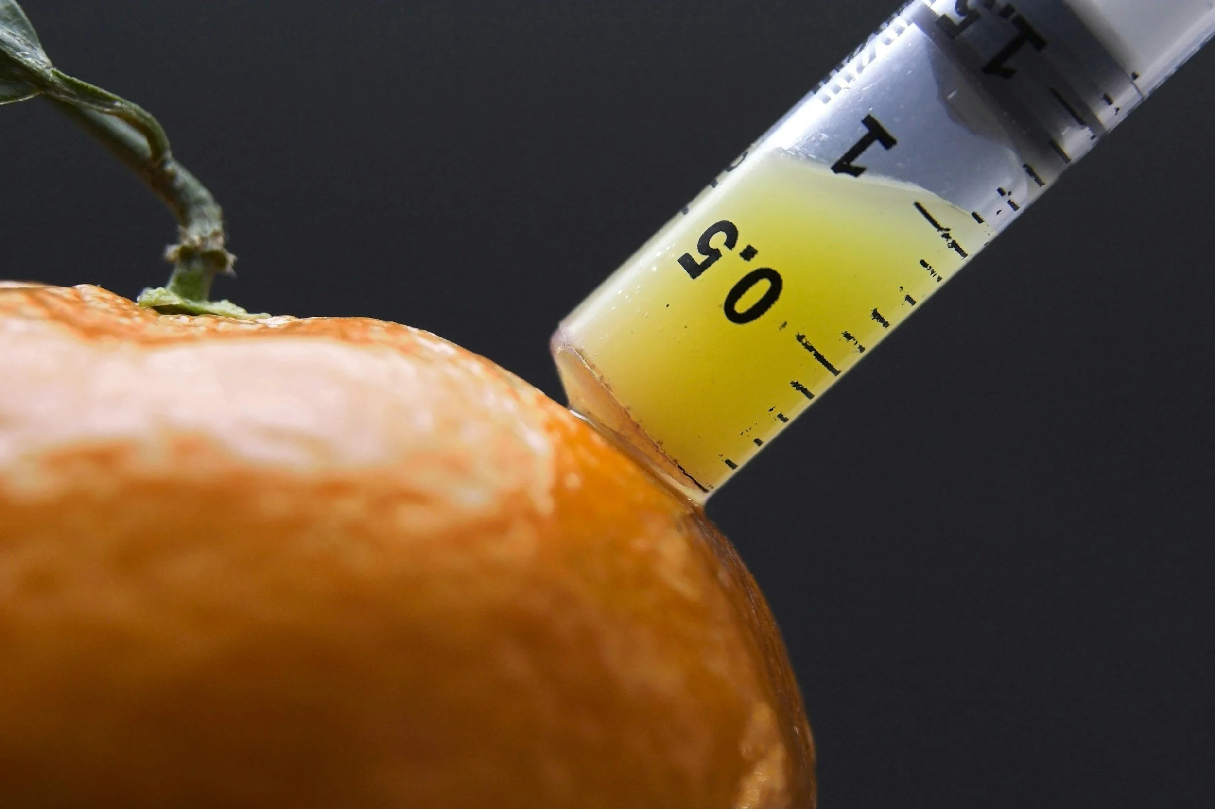 A close-up image of a syringe filled with yellow liquid, injecting into the skin of an orange fruit, with a dark background.