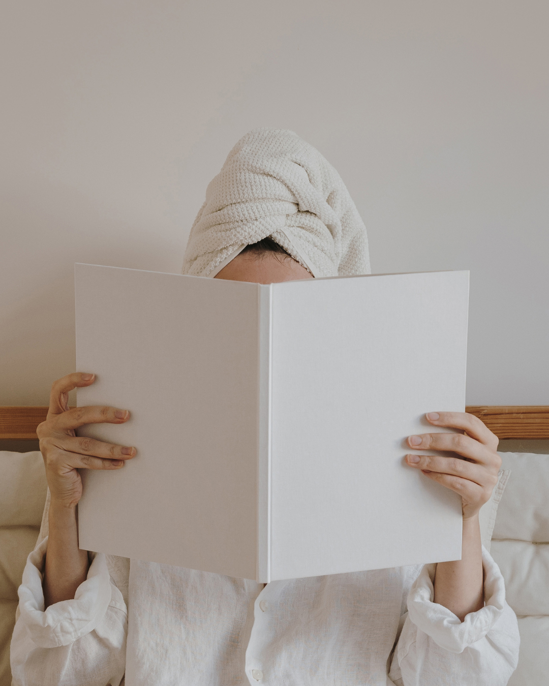 Person with a towel wrapped around their head reading a large white book or folder against a plain wall.