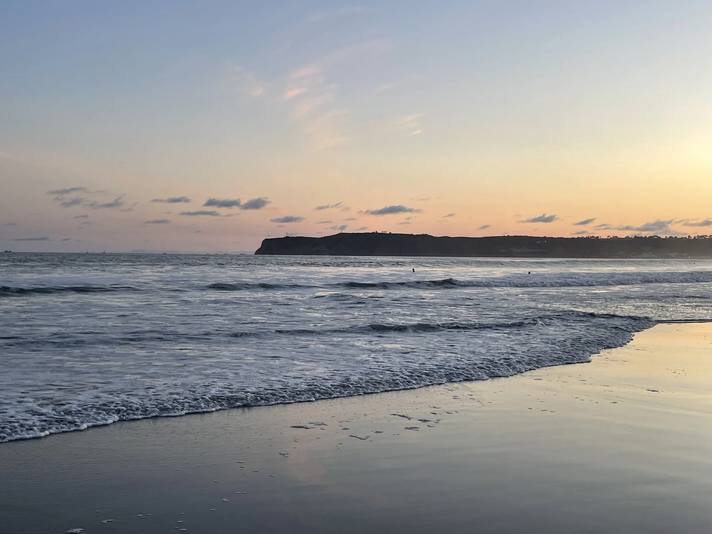 A serene beach scene at sunset with the sky painted in soft pastel colors, gentle waves washing onto the shore, and a dark silhouette of a cliff or headland in the background.