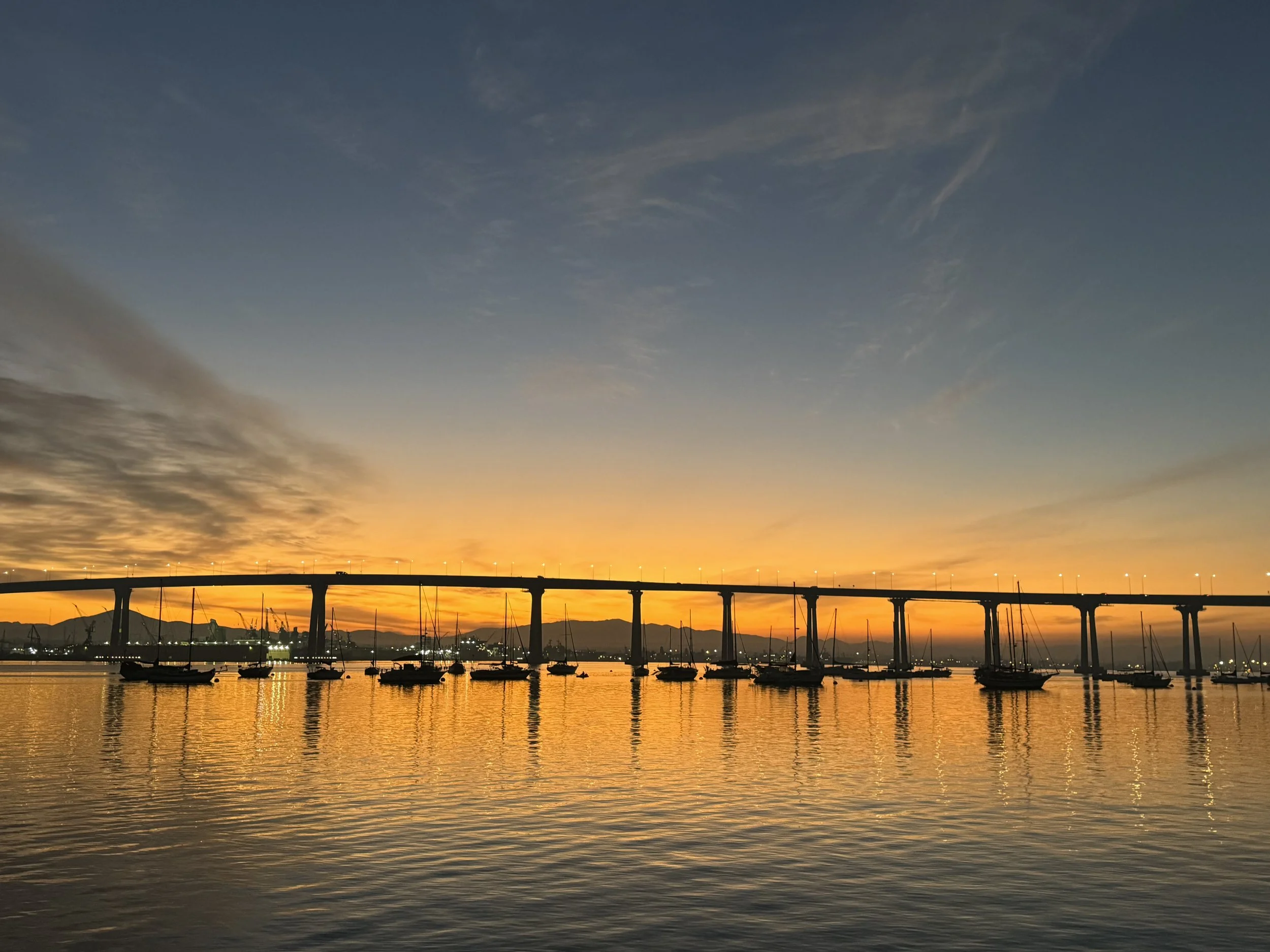 A sunset over a harbor with boats and a bridge, silhouetted against a sky with clouds and a gradient from orange to deep blue.