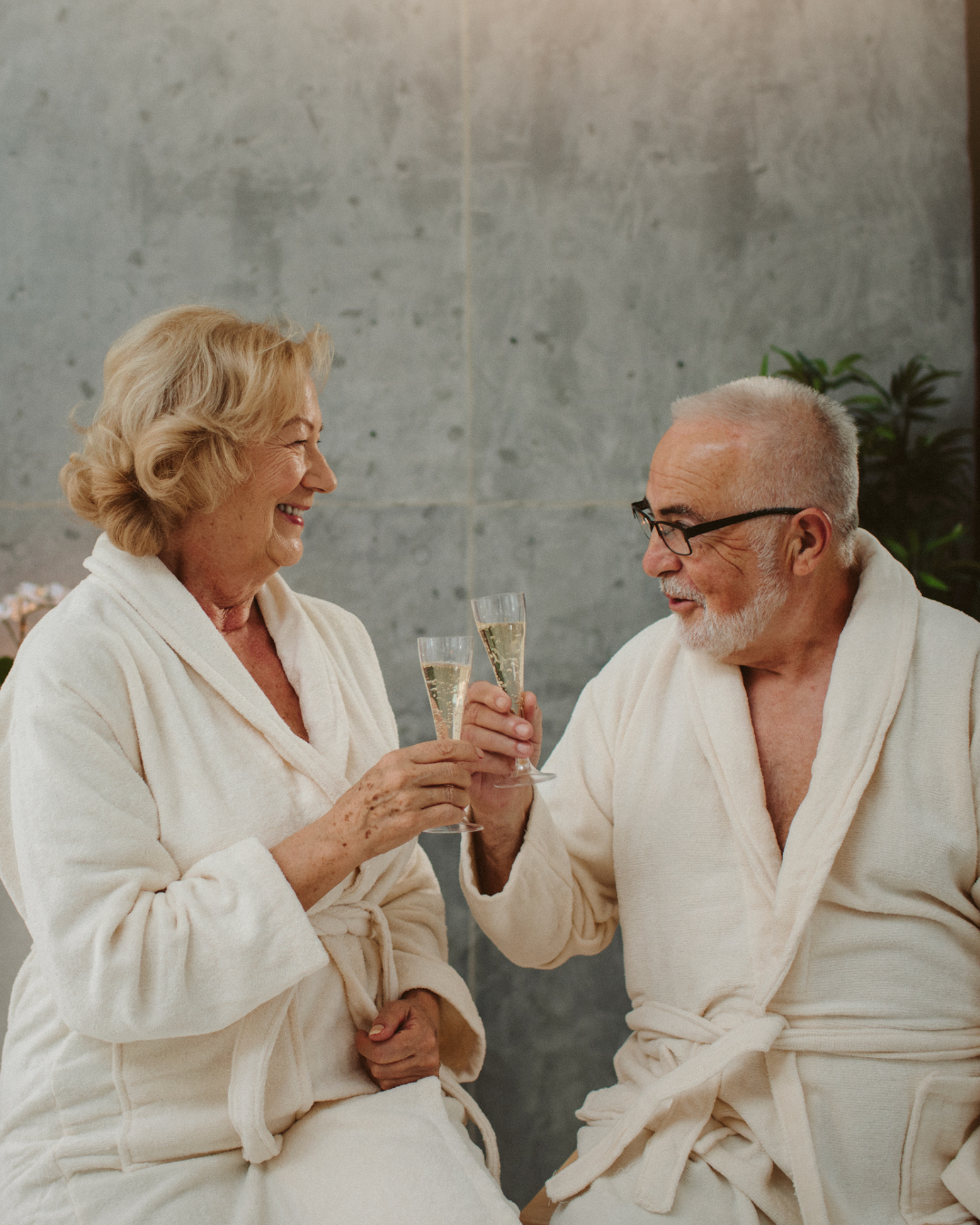 An elderly woman and man in bathrobes smiling and clinking glasses of champagne in a spa or wellness setting.