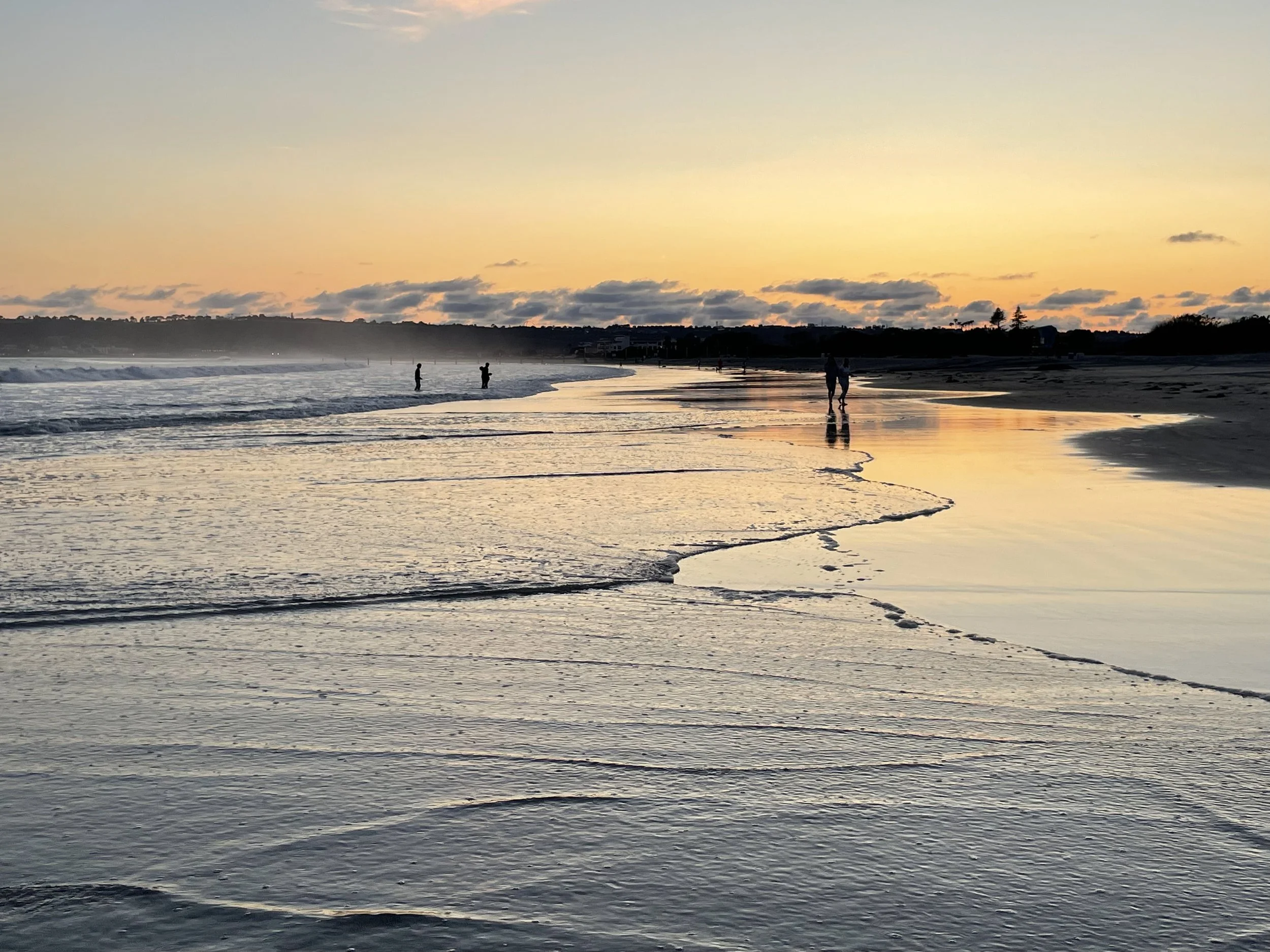 Sunset at the beach with calm waves and a few people walking along the shoreline, silhouetted against the colorful sky.