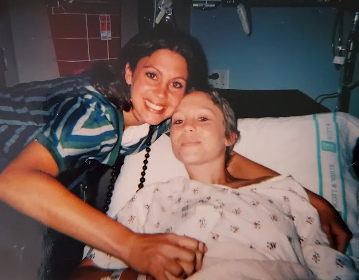 Patient Advocate Christi Jo poses next to her sister, who lies in a hospital bed, sick with cancer.