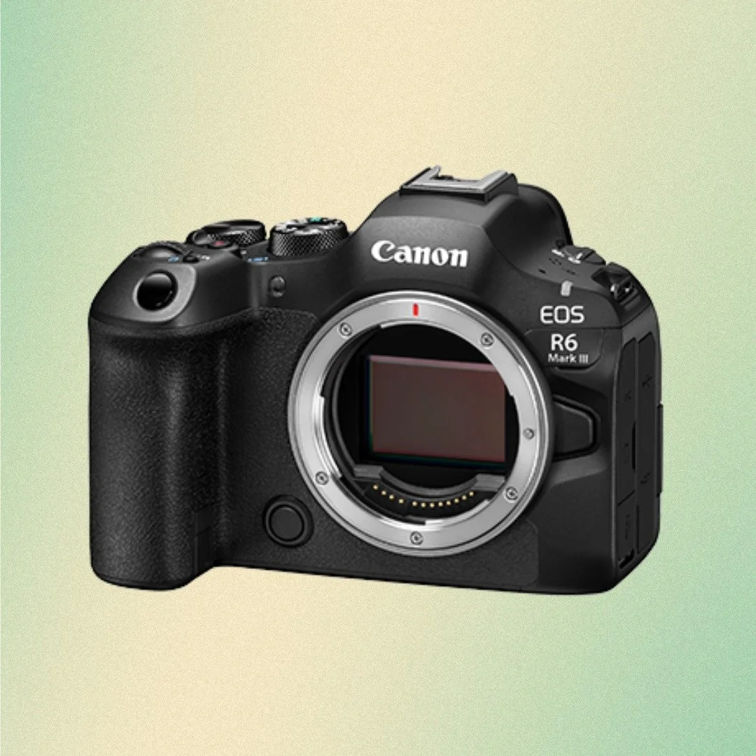 A person holding a Canon DSLR camera with a zoom lens in front of a city street with beige buildings, a parked car, trees, and cloudy sky.