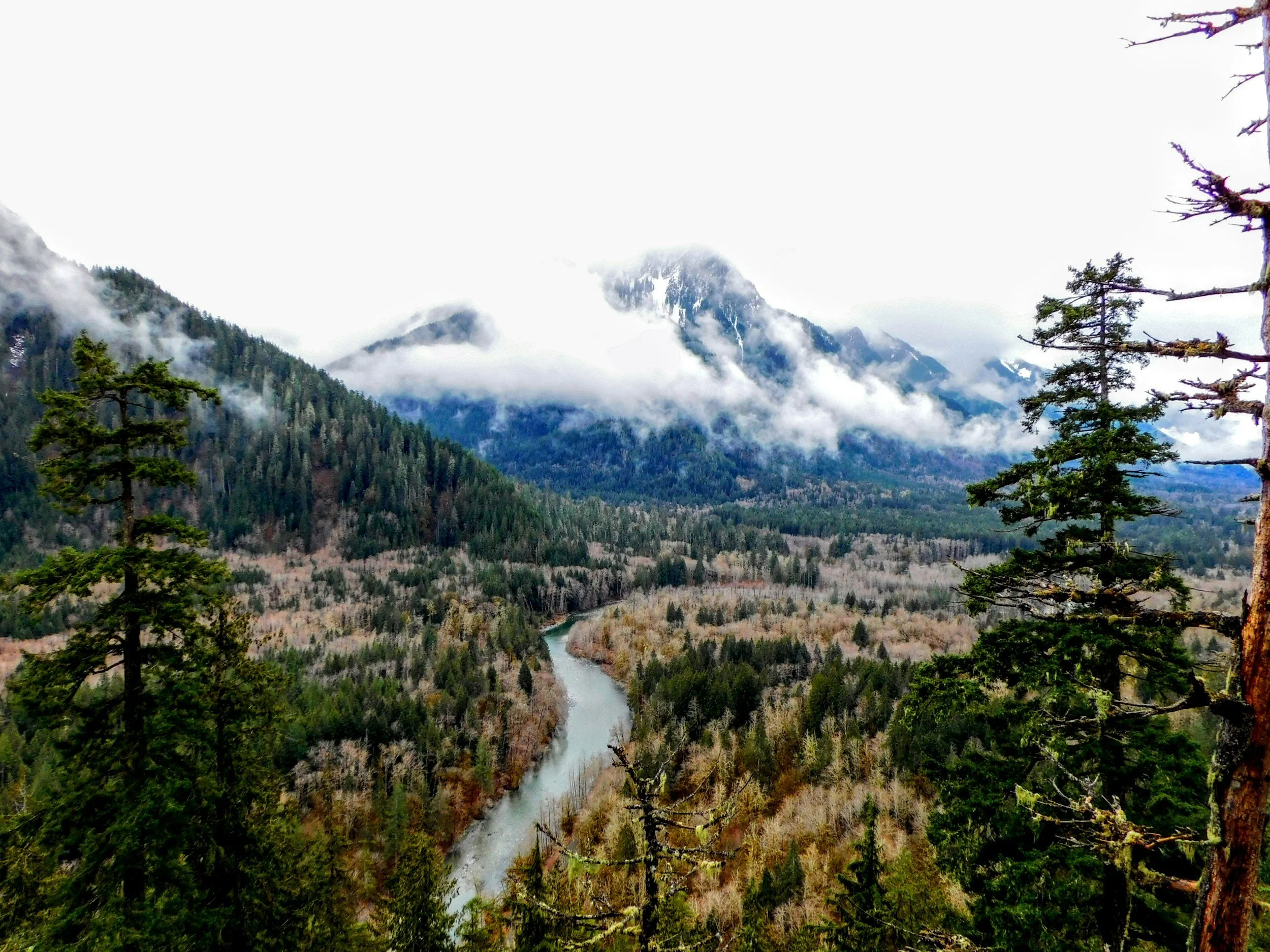 Mountain landscape with forested slopes, a river winding through the valley, and clouds surrounding the mountain peaks.