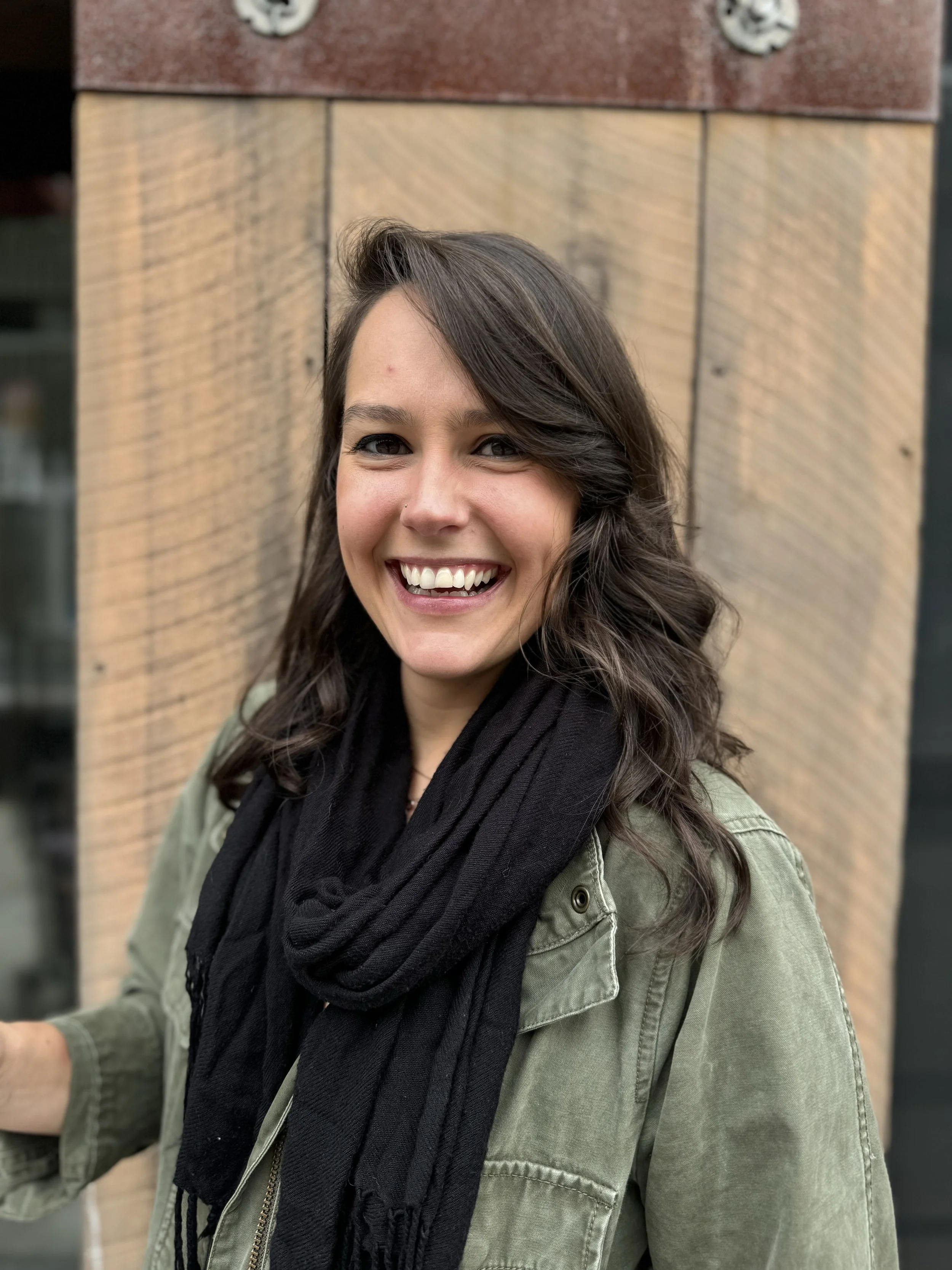 A young woman with dark hair styled in loose waves, smiling broadly, wearing a black scarf and a green military-style jacket with a zipper and button details, standing against a wooden wall background.